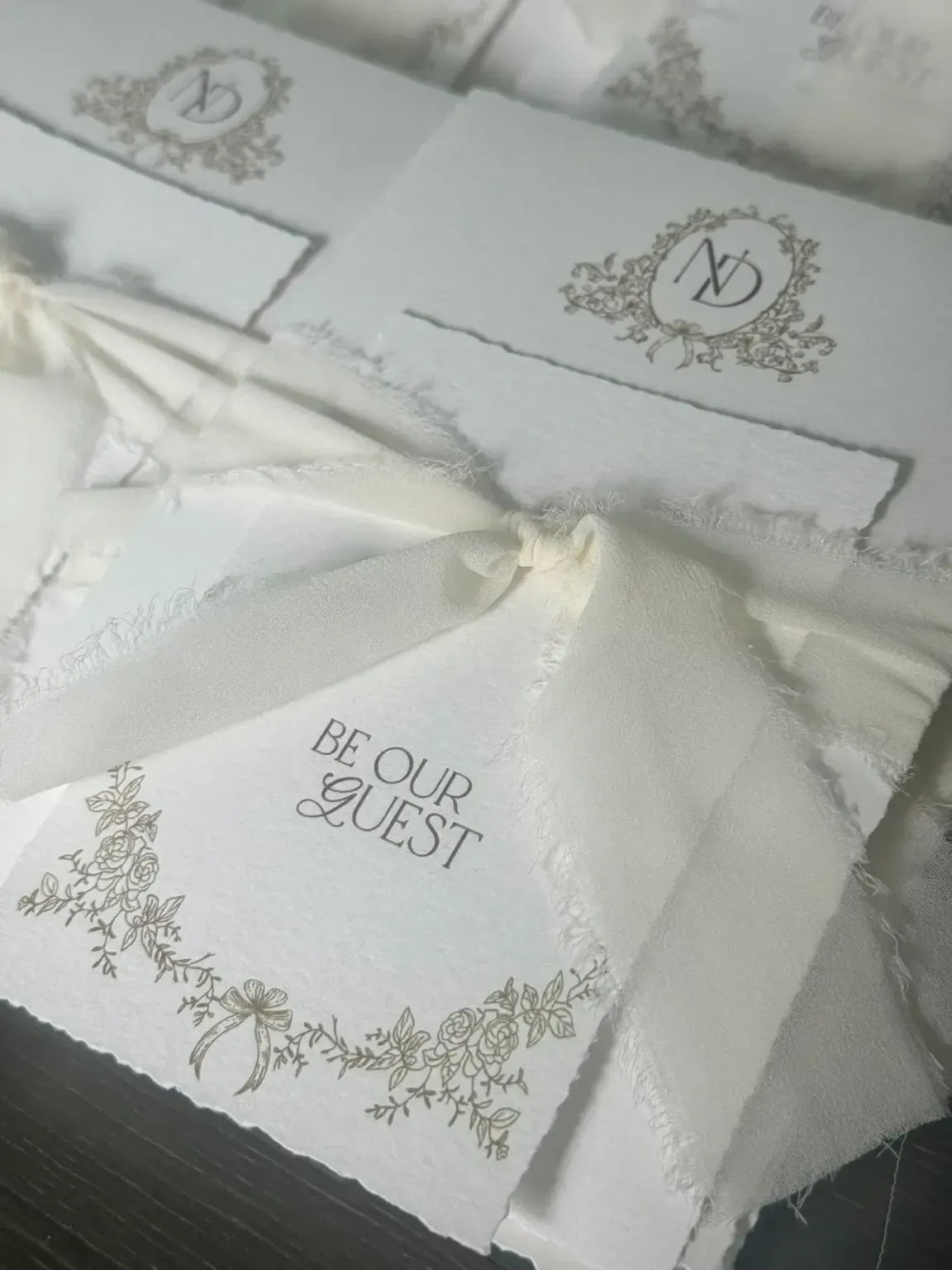 Cette image montre un ensemble d'invitations de mariage élégantes réalisées par Photogenia Design Studio, spécialiste des invitations de mariage. Les invitations sont ornées d'un motif floral délicat et d'un élégant ruban en tissu blanc attaché en nœud, ajoutant une touche sophistiquée. L'une des cartes porte l'inscription 'Be Our Guest' en lettres raffinées, suggérant une invitation à un événement nuptial distingué. Le papier arborant une finition texturée et des bords frangés, accentue l'artisanat de haute qualité.