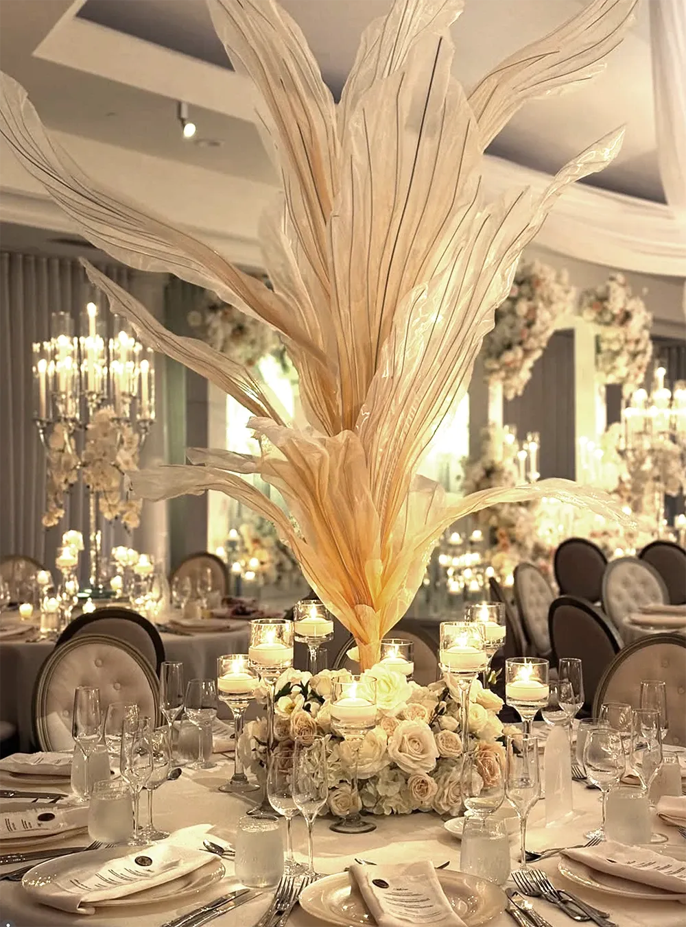 The image features a luxurious wedding reception setup by Zhen Decor, a vendor in the Wedding Decor category. At the center of a round table is an elaborate floral arrangement composed of large, elegant beige pampas grass plumes, complemented by an assortment of white roses and lush greenery. Surrounding the arrangement are several lit votive candles in glass holders, creating a warm, romantic ambiance. The table is elegantly set with fine china, crystal glasses, and neatly folded napkins atop pristine white tablecloths. In the background, tall, ornate candle holders with cascading flowers add to the sophisticated atmosphere of the venue, showcasing the artistry and attention to detail provided by Zhen Decor for weddings in Canada.