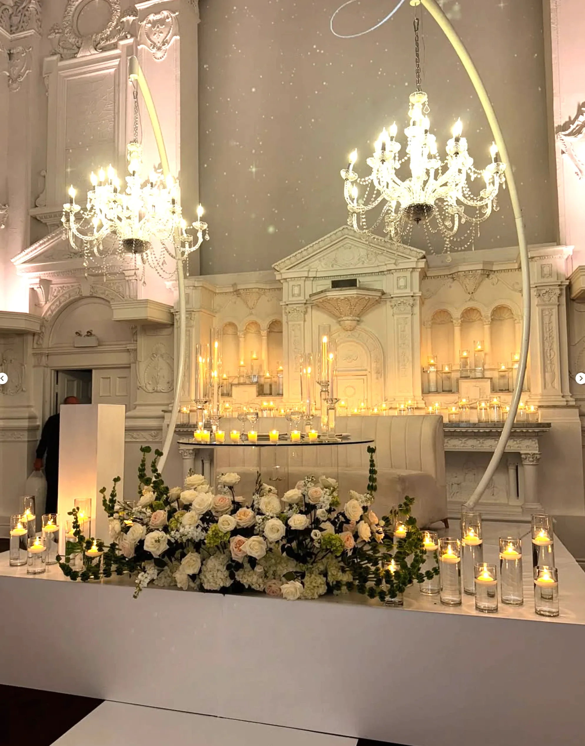 The image showcases an elegantly decorated wedding venue by Zhen Decor, a vendor in the Wedding Decor category at Mariage Elegant, Canada. The setting features an opulent arrangement of ivory and cream roses surrounded by lush greenery, beautifully accentuating a modern white altar. The backdrop is adorned with towering white columns and a grand classical arch, creating a majestic atmosphere. Dozens of glowing candles in clear glass holders illuminate the scene, casting a warm and inviting light. Extravagant chandeliers hang from the ceiling, adding a touch of luxury to the sophisticated ambiance.
