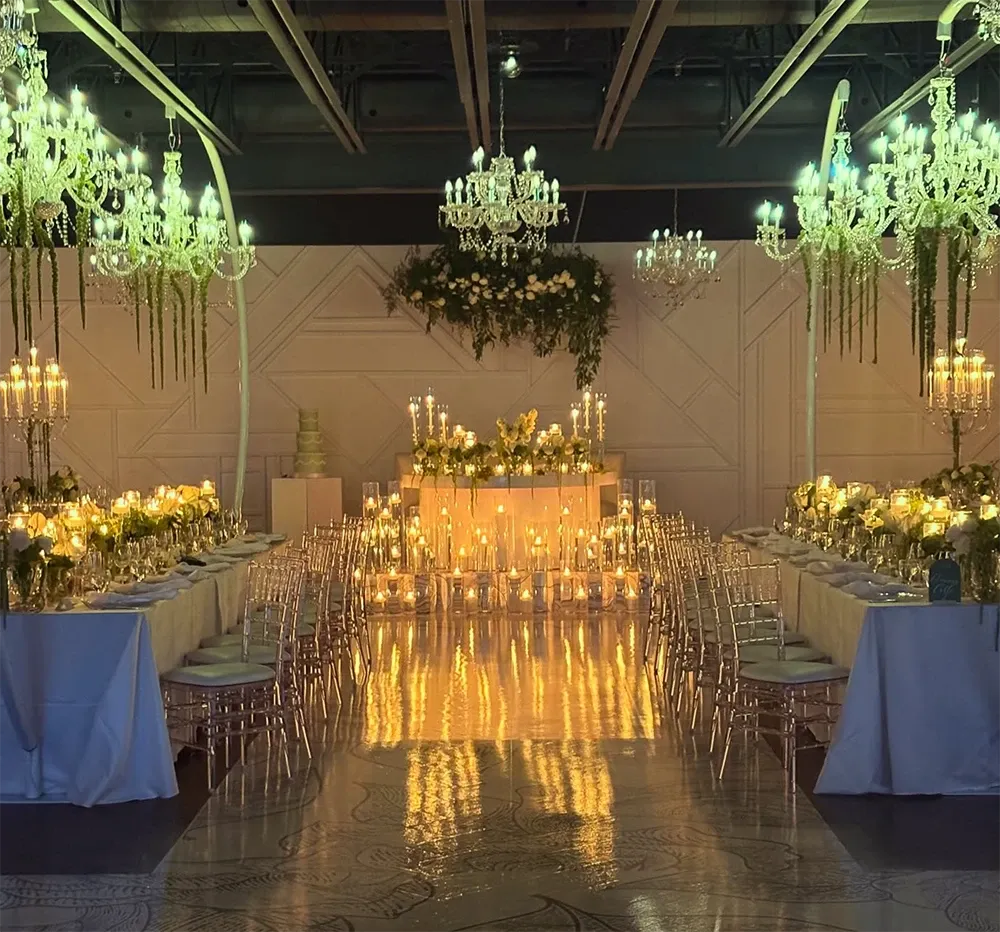 The image showcases a beautifully decorated wedding reception setup by Zhen Decor in the wedding decor category. The scene features two long banquet tables adorned with white tablecloths and elegantly arranged floral centerpieces, surrounded by clear, modern chairs. A central focus is a lavishly decorated head table, glowing with warm candlelight and enhanced by lush greenery. Elegant crystal chandeliers hang from the ceiling, casting a soft, romantic light over the venue. The ambiance is sophisticated and inviting, reflecting an attention to detail and style suitable for an elegant marriage event in Canada.