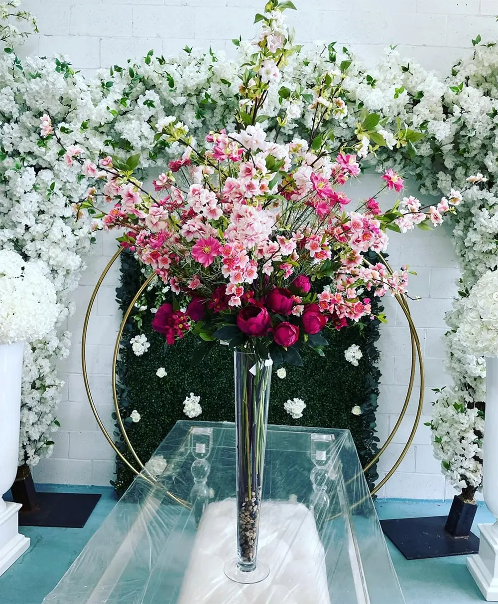 The image showcases an elegant wedding decor setup by Zhen Decor in the wedding decor category. A tall, clear vase overflowing with vibrant pink and white flowers, including peonies and cherry blossoms, commands attention at the center. Behind it, a lush, flower-covered wall with a backdrop of white blossoms elegantly frames a gold circular arch. The arrangement exudes sophistication, perfect for enhancing the aesthetic of a wedding ceremony or reception. White floral arrangements on pedestals flank the design, adding a symmetrical balance to the decor.