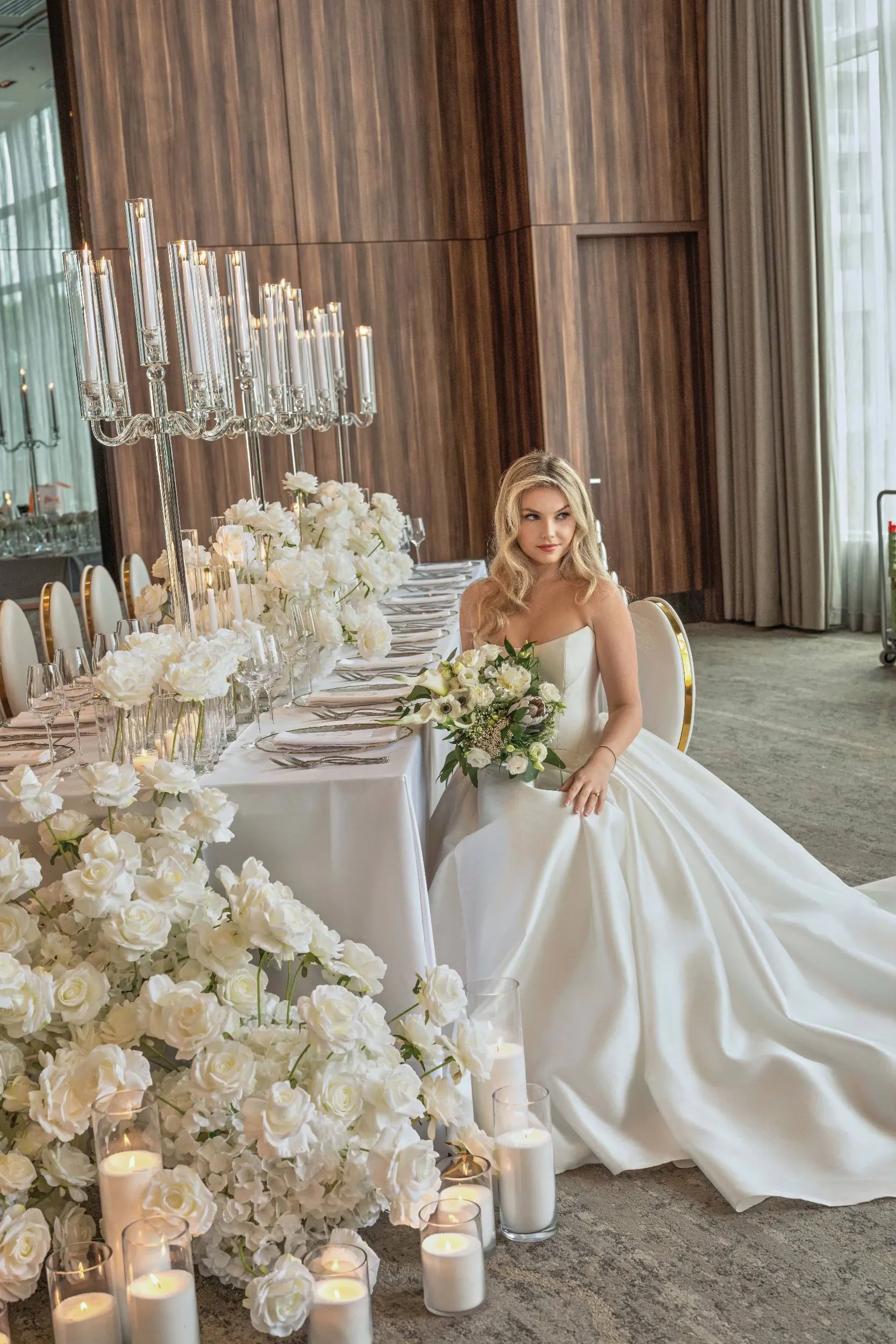 In the image, a bride in an elegant white gown is seated at a lavishly decorated wedding reception table. The decor, provided by Zhen Decor in the Wedding Decor category, features an abundance of white roses and greenery. A series of tall, slender glass candle holders line the center of the table, adding a touch of sophistication and romance to the scene. The setting is luxurious, with floor-to-ceiling wooden paneling and large windows letting in natural light. The table is set with fine china and cutlery, showcasing Zhen Decor's attention to detail and their expertise in creating a glamorous and stylish wedding atmosphere for Mariage Elegant in Canada.