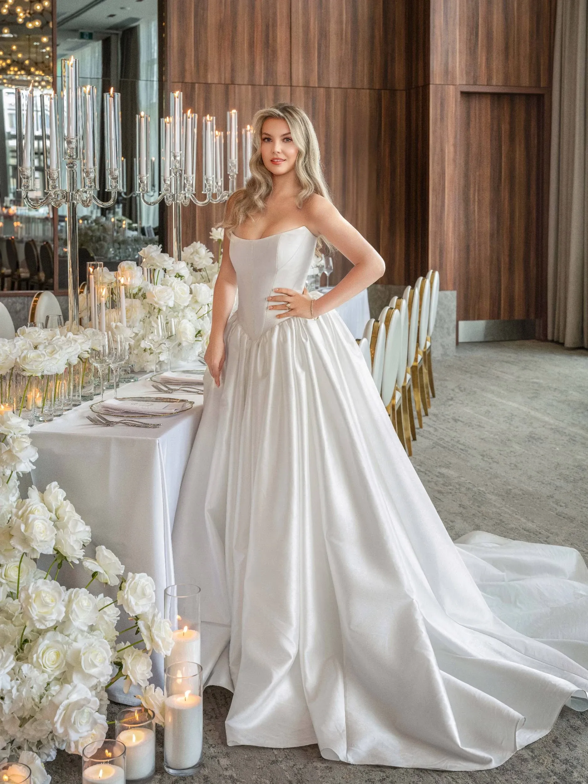 A bride in an elegant white gown stands beside a beautifully decorated wedding table by Zhen Decor, a wedding decor vendor featured on Mariage Elegant, Canada. The table is adorned with white floral arrangements and tall, silver candelabras, creating a luxurious and sophisticated atmosphere. White candles in glass holders are placed around the setting, enhancing the romantic ambiance. The background features wooden paneling and a mirror reflecting the soft lighting, adding warmth to the scene.