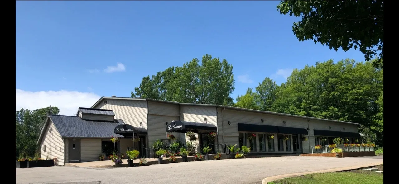 The image shows the exterior of Plaza du Boisé, a wedding venue listed on Mariage Elegant in Canada. The building is a large, single-story structure with a beige facade and a black roof. Several black awnings adorn the entrances, adding an elegant touch to the venue. The area in front of the building features well-maintained planters with green foliage and colorful flowers, enhancing its welcoming appeal. The venue is surrounded by lush green trees under a clear blue sky, providing a serene and picturesque backdrop for weddings and events.