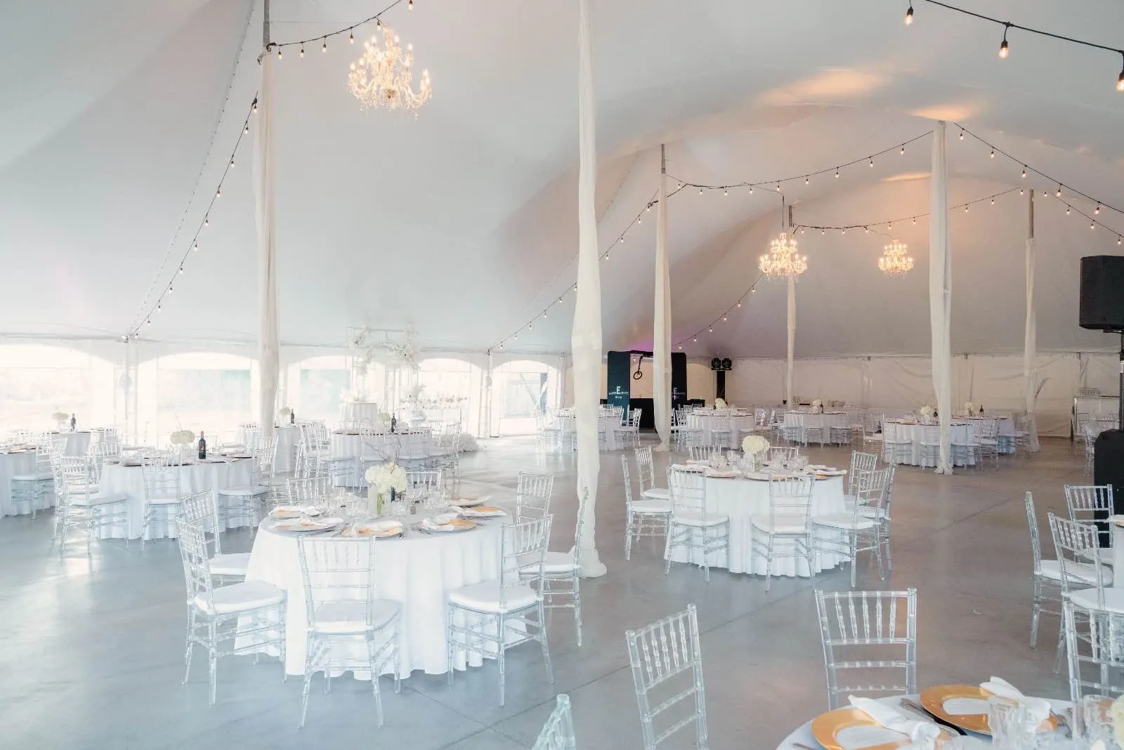 The image showcases Plaza du Boisé, a spacious wedding venue under the category of Wedding Venues, featured on Mariage Elegant, Canada. The elegant space is beautifully decorated, with an expansive white tented ceiling adorned with twinkling string lights and ornate chandeliers that exude a romantic ambiance. The floor plan is arranged with round tables dressed in pristine white linens, each elegantly set with gold-rimmed plates and neatly folded napkins. Clear, elegant chairs add a modern touch to the classic setup. Soft floral centerpieces grace the tables, enhancing the sophisticated decor, making it an ideal setting for weddings.