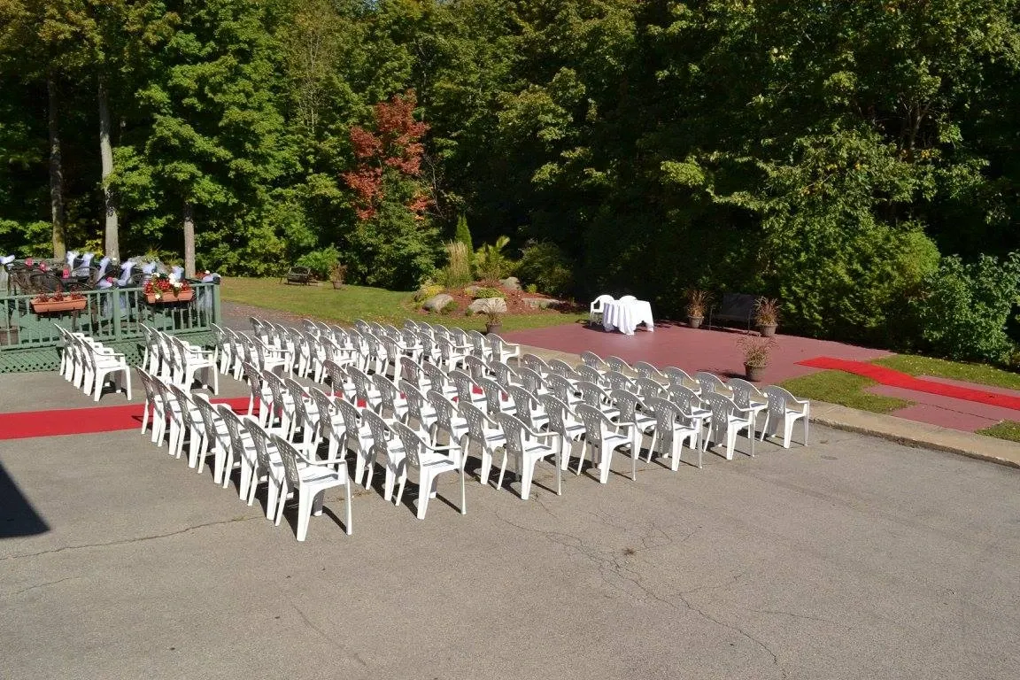 The image shows an outdoor wedding venue at Plaza du Boisé, categorized under Wedding Venues. Rows of neatly arranged white chairs are set up on a paved patio, facing an area intended for the ceremony, surrounded by lush green trees. A red carpet creates a walkway leading to a small stage or altar space, adding elegance to the natural setting. The peaceful forested backdrop provides a serene atmosphere, making it an ideal spot for outdoor weddings.