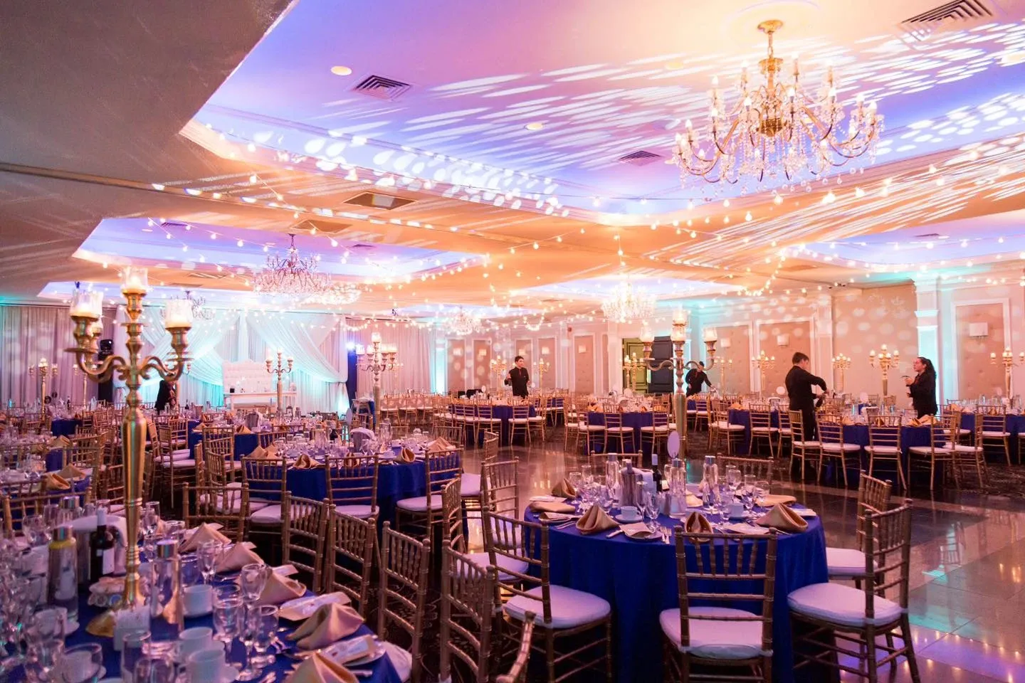 The image showcases the Plaza Universel, a wedding venue listed under the wedding planning and vendor site Mariage Elegant in Canada. The elegant hall is beautifully arranged for a wedding reception, featuring numerous round tables adorned with royal blue tablecloths and neatly folded napkins. Each table is set with sparkling glassware and fine china, reflecting the venue's attention to detail and sophistication. The ceiling is adorned with intricate chandeliers and adorned with twinkling string lights, casting a warm glow over the space. The ambiance is both luxurious and inviting, perfectly suited for a memorable wedding celebration. Attendees in the background contribute to the lively atmosphere, highlighting Plaza Universel as an ideal setting for special events.