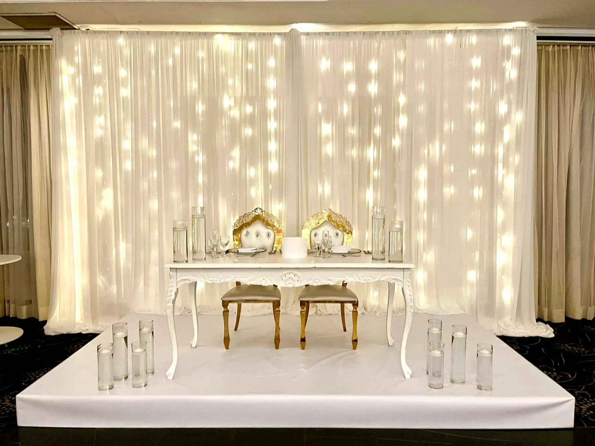 The image showcases a beautifully arranged wedding venue setup by Plaza Universel, a prominent vendor in the Wedding Venues category. The focal point is an elegant sweetheart table adorned with a pair of ornate, white chairs with gold detailing. The chairs are positioned behind a pristine white table, which is elegantly set with glassware and plates. The backdrop features a stunning curtain of delicate, warm fairy lights, casting a soft glow that enhances the romantic ambiance of the scene. The setting is accentuated by clear cylindrical vases placed around the platform, adding a sophisticated touch to the overall decor. This setup exemplifies the refined and luxurious wedding atmosphere offered by Plaza Universel in Canada.