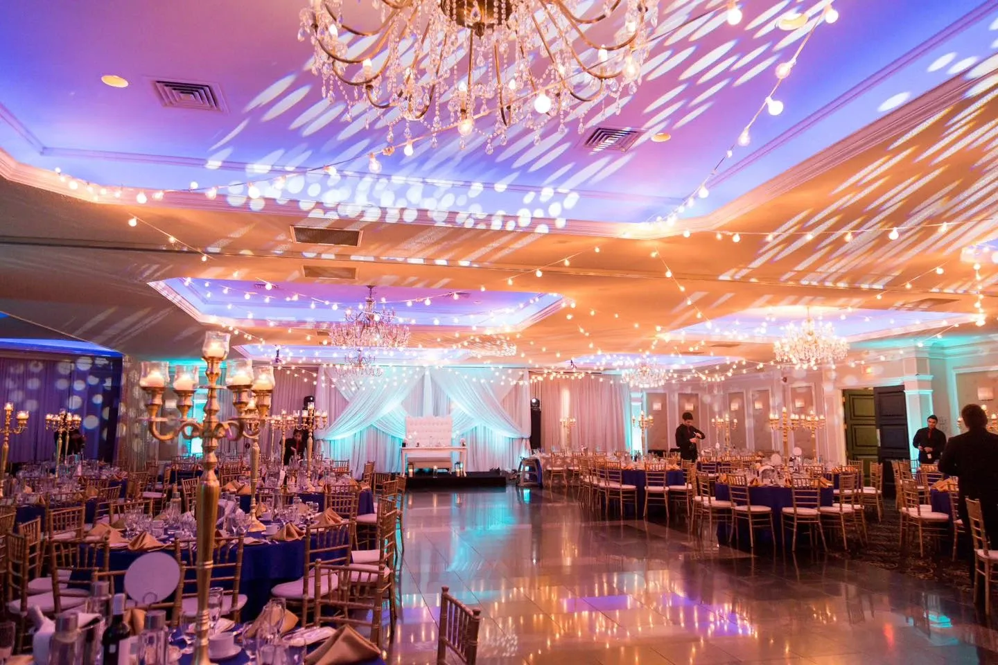 The image features an elegant wedding venue at Plaza Universel, showcasing its spacious and beautifully decorated interior. The category is Wedding Venues. The room is adorned with luxurious chandeliers and decorated with soft, ambient lighting that casts a romantic glow. Tables are elegantly set with fine tableware and surrounded by gold chiavari chairs. The ceiling is draped with string lights, adding a whimsical atmosphere to the setting. A backdrop, likely for the wedding party, is situated at the end of the room, framed by soft white drapery and accented by gentle blue uplighting. The polished floor reflects the vibrant lights above, contributing to the overall stylish and sophisticated ambiance perfect for wedding celebrations.