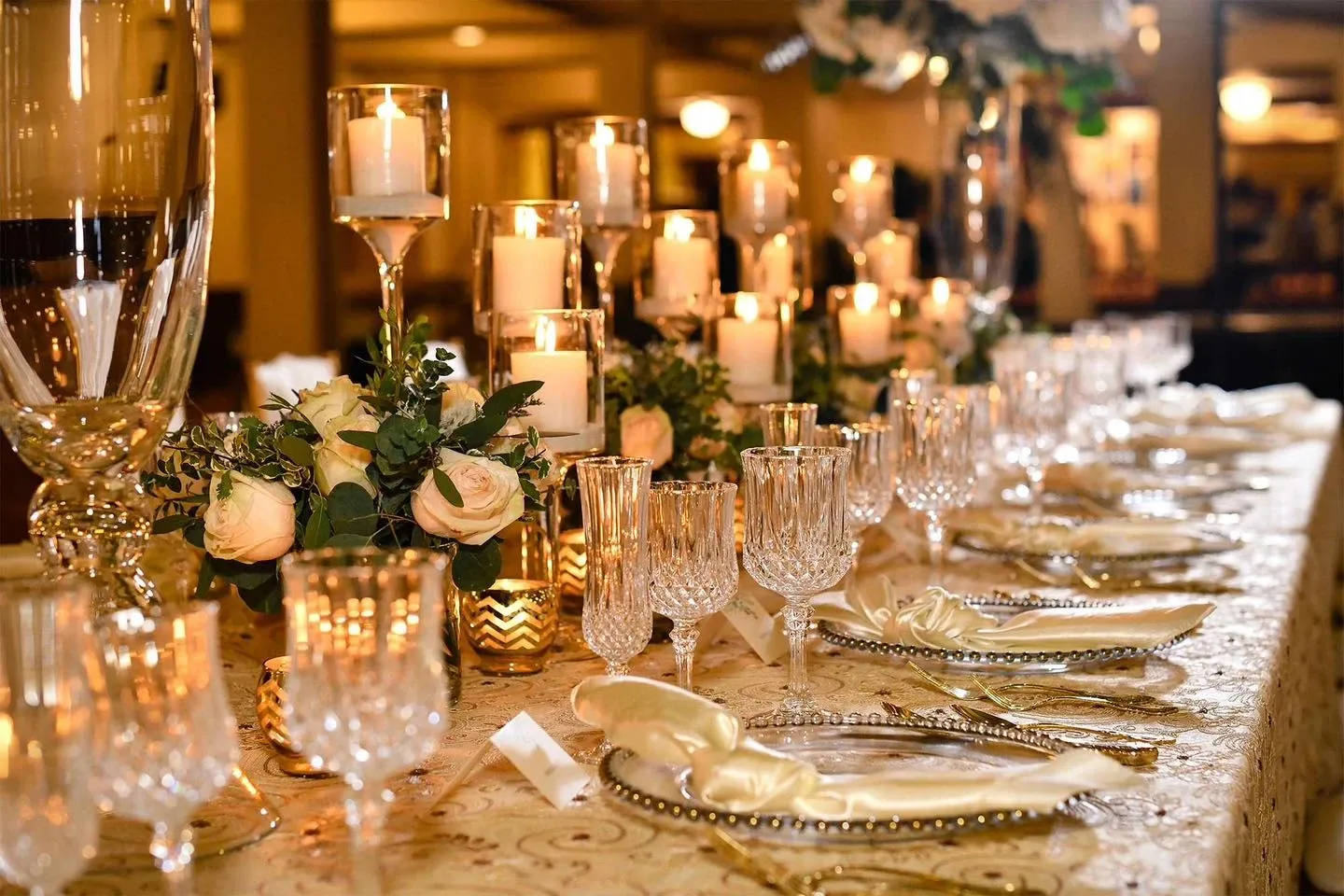This image showcases an elegant wedding reception setup at Plaza Universel, a renowned wedding venue. The table is adorned with an exquisite arrangement of white roses and lush greenery, complemented by a series of tall, glowing candles that create a warm, inviting ambiance. Crystal glassware and intricately designed plates, embellished with delicate gold elements, are meticulously arranged, enhancing the luxurious feel of the setting. This refined display highlights Plaza Universel’s attention to detail and sophistication, making it an ideal choice for couples planning their dream wedding at a premier venue in Canada.