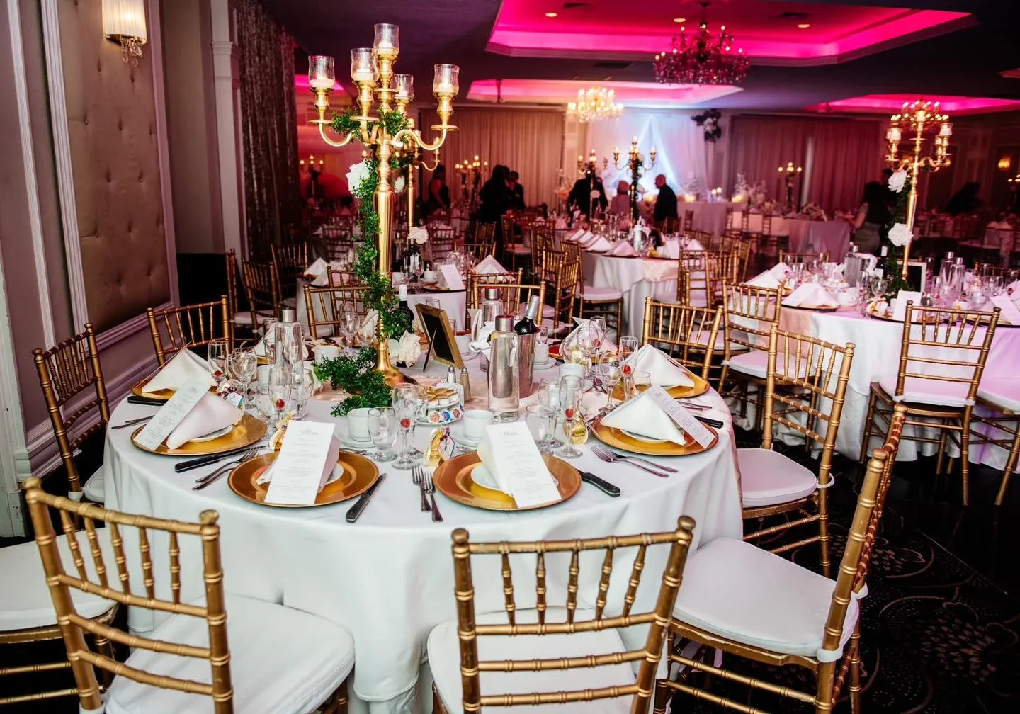 The image showcases a beautifully decorated wedding venue at Plaza Universel, a featured vendor in the Wedding Venues category on Mariage Elegant, Canada. The elegant setting includes round tables covered with crisp white linens, each adorned with gold-rimmed plates, neatly folded napkins, and detailed menus. Golden Chiavari chairs encircle the tables, enhancing the sophisticated atmosphere. The centerpiece features ornate, gold candelabras with flickering candles, adding a romantic glow to the venue. The background displays other decorated tables with similar setups and warm ambient lighting, contributing to the overall luxurious and inviting environment for a wedding celebration.