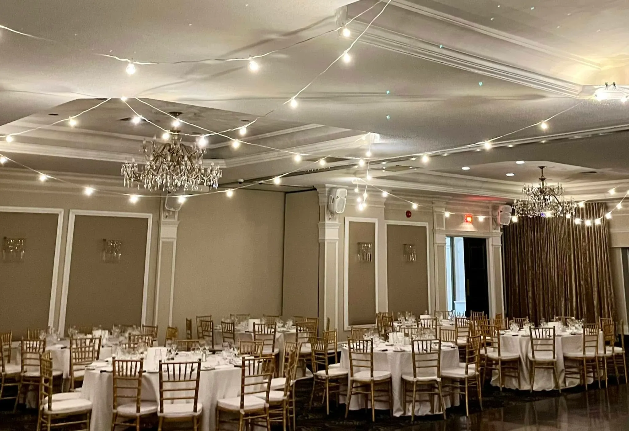 The image shows an elegantly arranged wedding venue at Plaza Universel under the wedding venues category. The spacious room features tables covered with crisp white tablecloths, each surrounded by gold Chiavari chairs. Soft, ambient lighting emanates from strings of delicate lights and ornate chandeliers hanging from the ceiling. The venue's neutral-toned walls are accented with intricate molding, creating a sophisticated and inviting atmosphere for wedding celebrations.