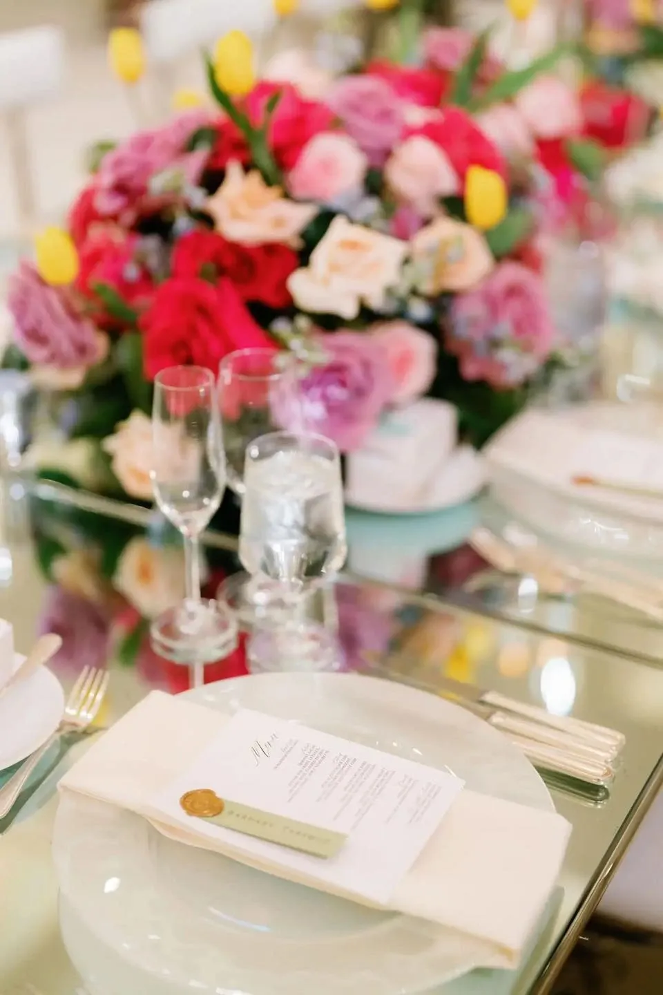 The image showcases a beautifully set wedding reception table at Plaza Universel, a premier wedding venue in Canada. The table features elegant clear glassware and polished silver cutlery. The centerpiece is an exquisite floral arrangement comprised of vibrant red, pink, and yellow flowers, adding a pop of color to the setup. Delicate white plates are placed at each setting, with a neatly folded napkin and a sophisticated menu card on top, enhancing the luxurious ambiance of the venue. This setup is perfect for couples seeking an elegant and refined atmosphere for their special day.