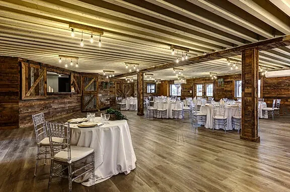 The image showcases the interior of Plaza Rive Nord, a wedding venue in the category of Wedding Venues listed on Mariage Elegant, Canada. The spacious room features rustic wooden walls and beams, giving it a warm, inviting atmosphere. Elegant round tables are set with white tablecloths, neatly arranged with place settings and decorative items. Chiavari chairs add a touch of sophistication, complementing the room's natural tones. Overhead, string lighting casts a soft glow, enhancing the venue's charm, perfect for hosting romantic wedding celebrations.