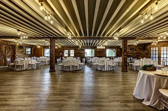 The image shows an elegant wedding venue at Plaza Rive Nord, categorized under wedding venues. The spacious room features rustic wooden beams and paneling, creating a warm, inviting atmosphere. Round tables are beautifully set with white tablecloths and elegantly draped chairs, ready for a wedding reception. Soft overhead lighting adds a romantic glow, highlighting the venue's charm. The room's large windows offer natural light, enhancing the sophisticated ambiance, ideal for weddings and special celebrations.