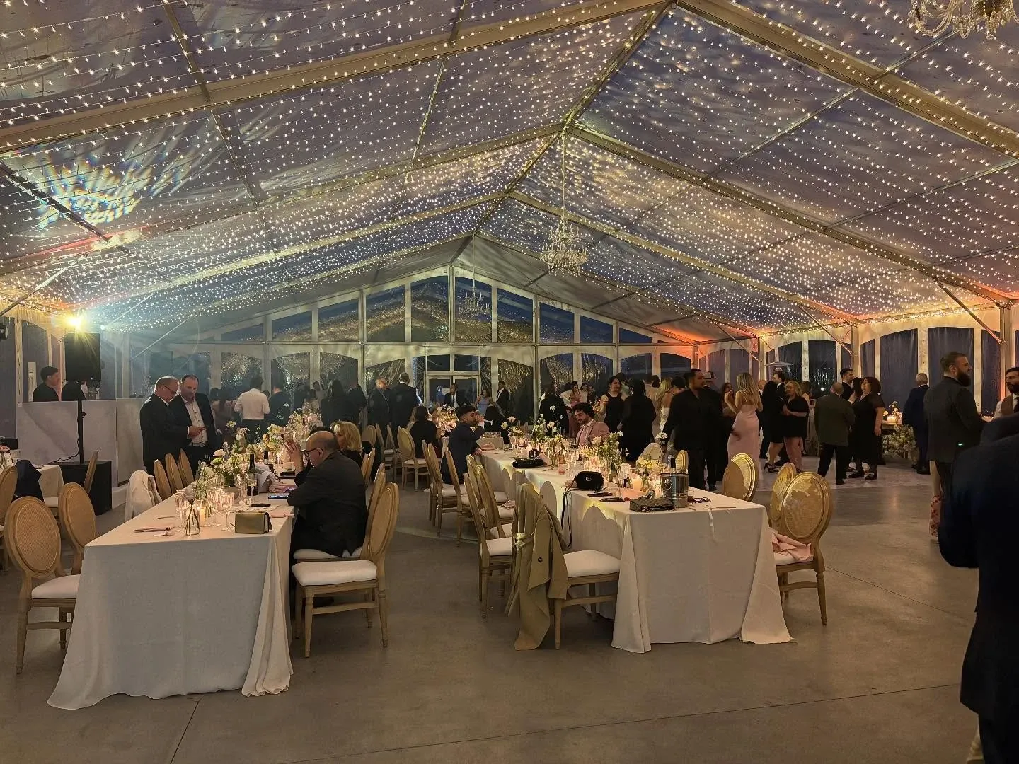 The image shows an elegant wedding event at Plaza Rive Nord, a venue featured under the wedding category on Mariage Elegant, Canada. The scene captures a spacious, transparent tent illuminated by strings of twinkling lights that create a romantic ambiance. Inside, round tables with white tablecloths are elegantly set with floral centerpieces and surrounded by cushioned chairs. Guests are mingling and enjoying the celebration, contributing to the lively and joyous atmosphere. The venue’s sophisticated setup highlights its suitability for hosting upscale wedding receptions.