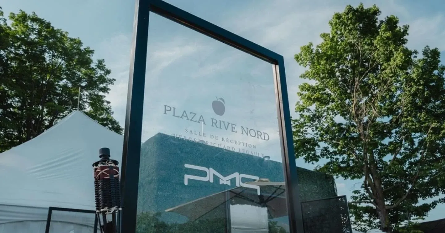 The image features the exterior of Plaza Rive Nord, a prominent wedding venue in Canada, part of the Mariage Elegant vendor listing. Dominating the foreground is a large, clear glass sign displaying the name 'Plaza Rive Nord' with additional text underneath that seems related to the venue’s offerings. In the background are lush green trees and part of a white tent, suggesting a picturesque and serene outdoor setting ideal for weddings. The setup hints at an elegant and inviting atmosphere, perfect for hosting wedding receptions and ceremonies.