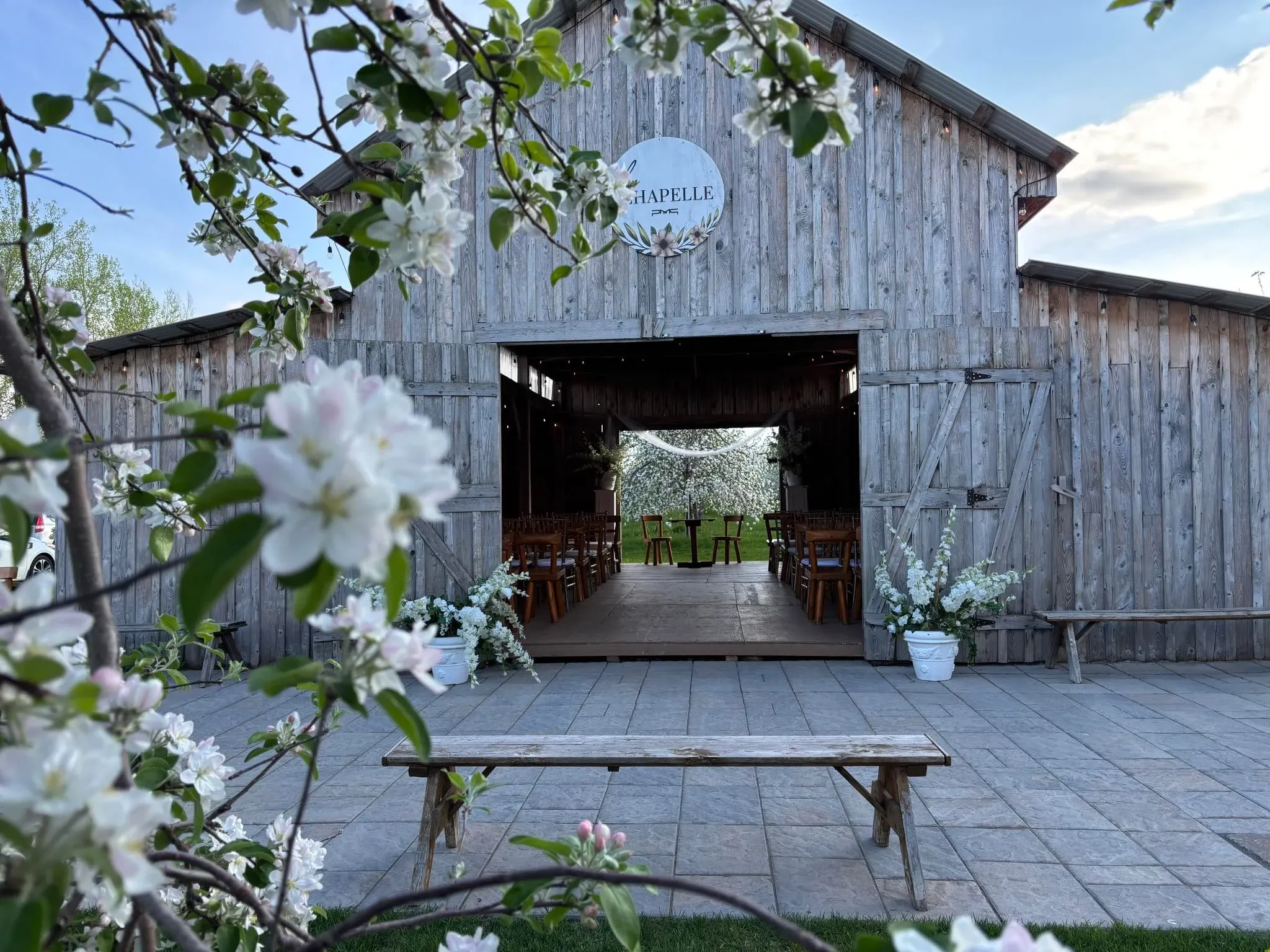 The image features Plaza Rive Nord, a wedding venue located in Canada, under the Mariage Elegant site. The photograph captures a rustic wooden barn adorned with elegant floral arrangements, perfect for a charming countryside wedding. The barn doors are open, revealing wooden chairs neatly arranged inside. In the foreground, blooming branches frame the scene, adding a touch of natural beauty. A wooden bench sits on a stone pathway leading up to the barn, enhancing the venue's rustic charm. This idyllic setting makes Plaza Rive Nord an appealing choice for couples seeking a romantic venue for their special day.