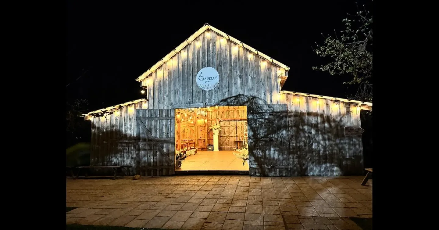 The image shows the exterior of Plaza Rive Nord, a charming wedding venue under the category of Wedding Venues on Mariage Elegant, Canada. The building has a rustic wooden facade, elegantly decorated with warm string lights along the roofline, creating a welcoming ambiance. The entrance is open, revealing a softly lit interior with wooden beams and floral arrangements, hinting at the venue's suitability for cozy and intimate wedding celebrations. Shadows of tree branches add a natural, enchanting element to the scene.