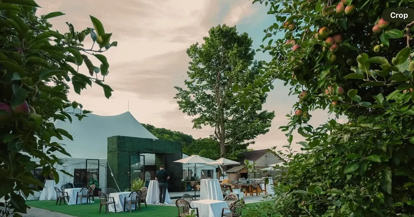 The image features the picturesque outdoor wedding venue, Plaza Rive Nord, categorized under Wedding Venues for Mariage Elegant in Canada. The venue is beautifully set amid lush greenery, with a large, elegant white canopy tent providing a shaded area. Underneath, round tables with white tablecloths are neatly arranged, ready for guests. Chairs are stylishly placed around the tables, and a well-maintained lawn enhances the overall look. In the background, a tree adds a natural touch to the scene, and apple trees on either side frame the venue beautifully, making it a charming setting for a wedding ceremony or reception.