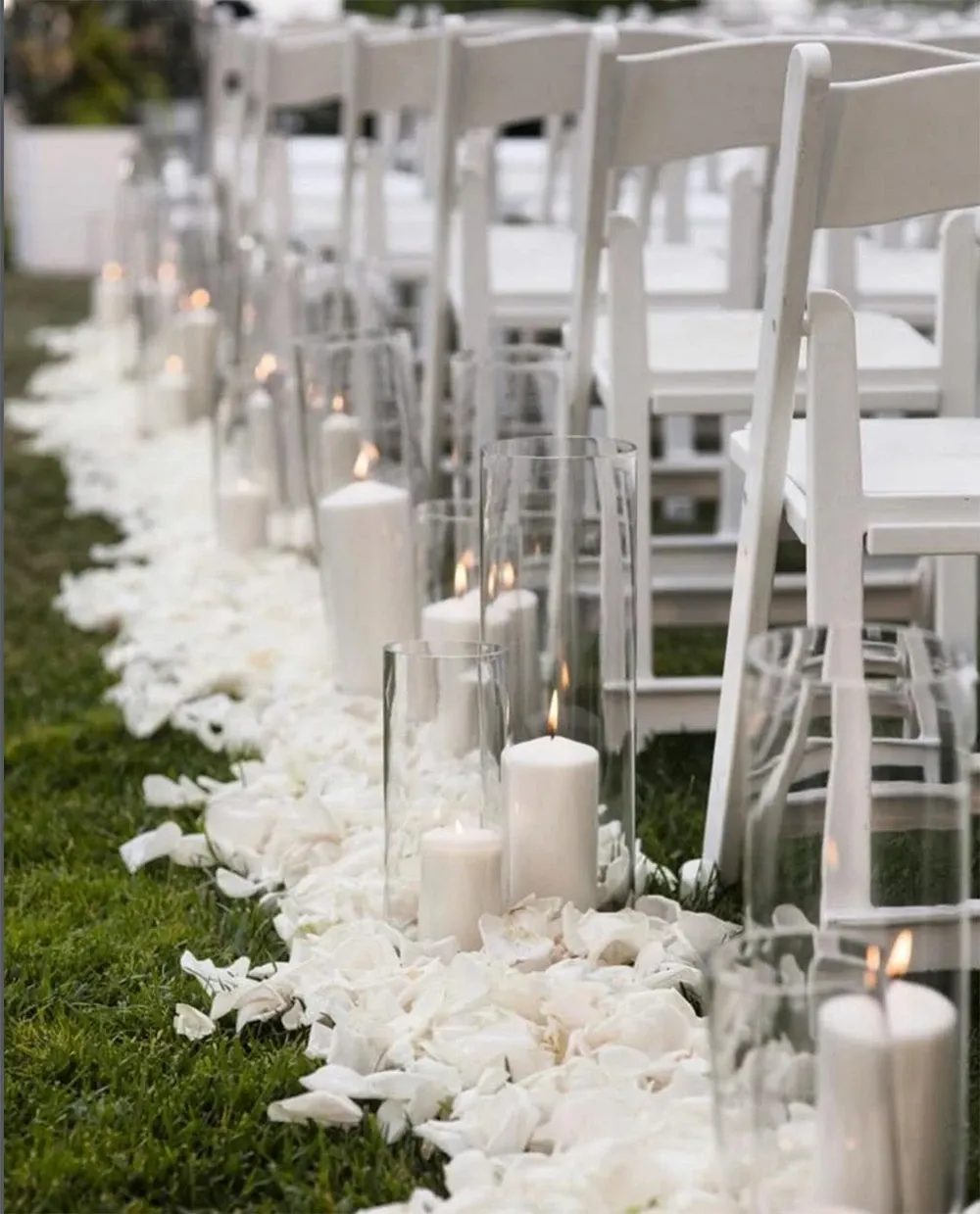 The image features an elegant wedding ceremony setup by Vizion, a wedding planner vendor in the Wedding Planners category at Mariage Elegant, Canada. It shows a row of white folding chairs aligned on a grassy aisle adorned with a lush layer of white rose petals. Tall glass cylinders holding white pillar candles are placed along the aisle, adding a romantic and sophisticated ambiance. The soft candlelight creates a warm and inviting atmosphere, perfect for a luxurious outdoor wedding celebration.