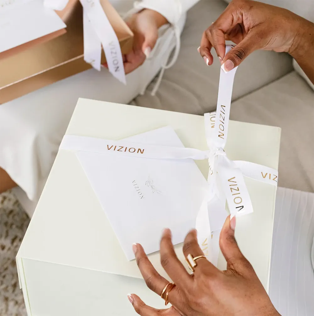 A close-up image featuring elegantly wrapped wedding gifts, showcasing the expertise of Vizion, a top vendor in the Wedding Planners category. The image displays a person’s hands delicately tying a ribbon on a sophisticated white gift box, adorned with custom ribbons inscribed with 'VIZION.' A white envelope, presumably containing a wedding-related message or invitation, rests atop the box. Another similar gift box is partially visible nearby, hinting at the detailed planning and preparation involved, emblematic of Vizion's commitment to creating memorable wedding experiences, as featured on Mariage Elegant, Canada.