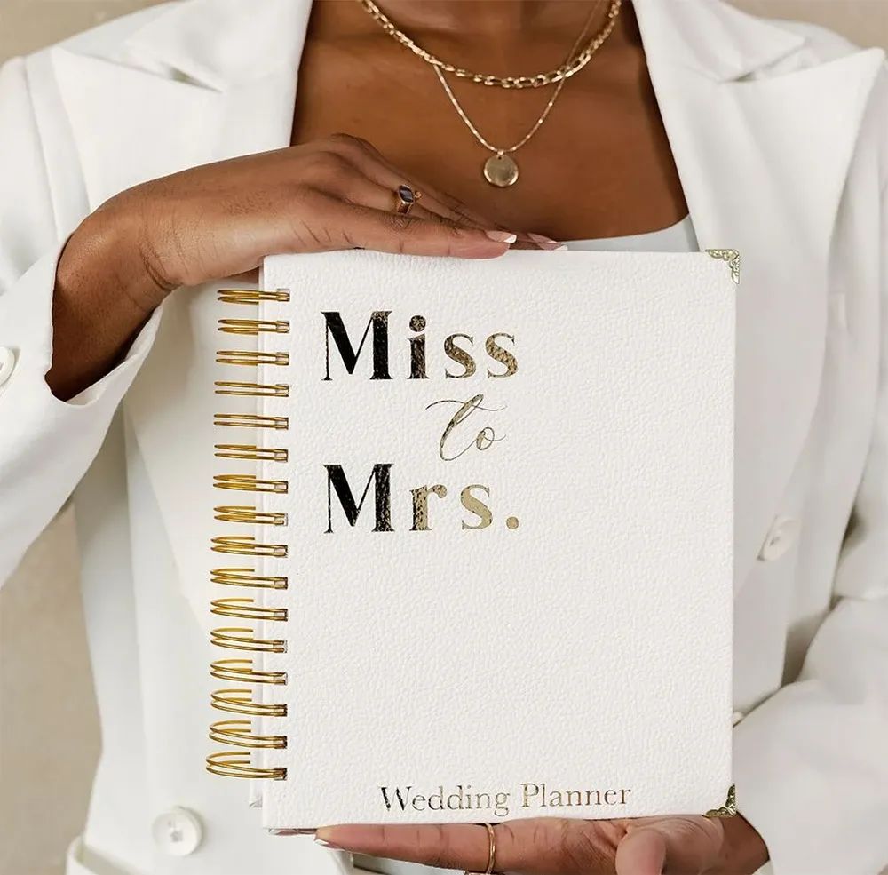 A woman is holding a stylish white wedding planner book embossed with the words 'Miss to Mrs.' in elegant gold lettering. Below, 'Wedding Planner' is subtly imprinted. She is dressed in a sophisticated white blazer and layered gold necklaces, adding a touch of elegance. This image represents Vizion, a distinguished name in the Wedding Planners category on Mariage Elegant in Canada, highlighting their attention to detail and commitment to creating memorable wedding experiences.