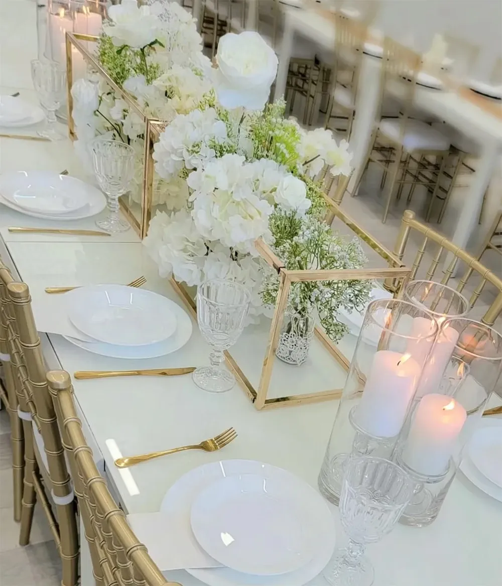 This image showcases an elegant table setting designed by the wedding planner Vizion, categorized under Wedding Planners for Mariage Elegant in Canada. The table features a sophisticated centerpiece with lush white flowers, including roses and hydrangeas, arranged in a modern gold geometric frame. Surrounding the floral display are several crystal candlesticks with tall white candles, adding a romantic ambiance. The table is set with white plates and glassware, complemented by gold cutlery. Beige or champagne-colored chiavari chairs are visible in the background, enhancing the overall tasteful and upscale wedding atmosphere.