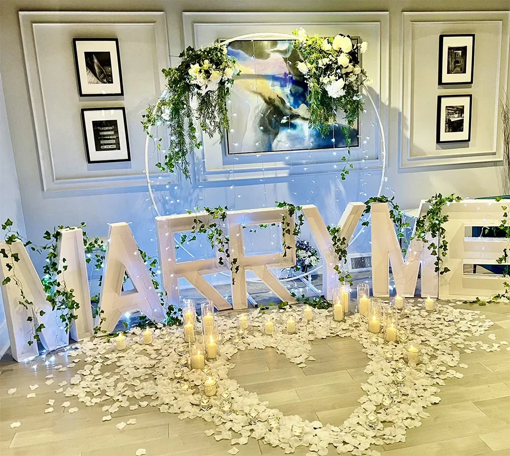 The image showcases a romantic wedding proposal setup designed by Vizion, a wedding planner featured in the Mariage Elegant vendor listing for Canada. The scene includes large, illuminated letters spelling 'MARRY ME,' adorned with cascading green ivy. In front of the letters, numerous candles are arranged surrounding a heart shape outlined with white rose petals. Above the proposal display, elegant floral arrangements featuring white roses and greenery add a graceful touch to the setting. The background features framed art on a subtly textured wall, enhancing the sophisticated ambiance.