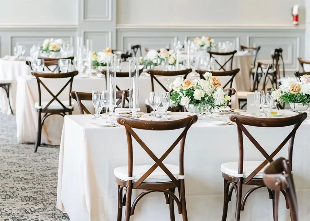 The image shows an elegantly arranged wedding reception setup, featuring tables adorned with white tablecloths and beautifully arranged floral centerpieces of white and peach roses. Each table is complemented by stylish wooden chairs with intricate cross-back designs. The setting, provided by the wedding planners Vizion, reflects sophistication and attention to detail, ideal for creating a romantic and memorable wedding atmosphere. The soft lighting and well-coordinated decor elements add to the ambiance, making it a perfect example of Vizion's expertise in the wedding planning category. The background showcases light grey paneling, enhancing the venue's elegant aesthetic.