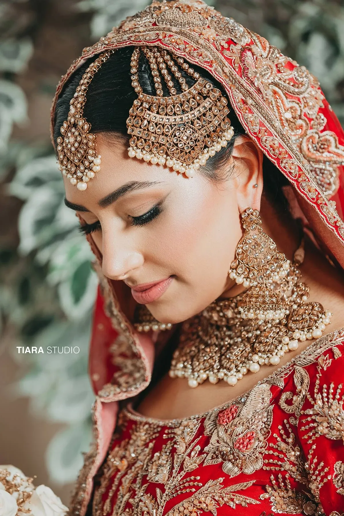 This image from Tiara Studio, a wedding photography vendor listed on Mariage Elegant in Canada, features a bride in a richly embroidered red and gold traditional wedding outfit. She is adorned with elaborate gold and pearl jewelry, including a striking maang tikka, large earrings, and an intricate necklace. The bride's eyes are gently closed, showcasing her elegant makeup. A soft focus background with greenery enhances the elegance of the portrait.