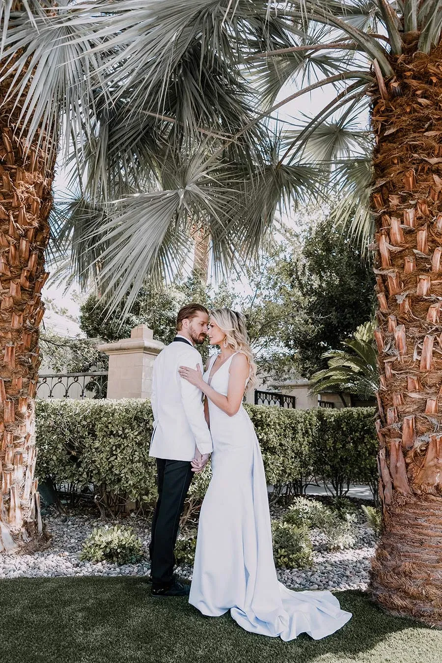 A bride and groom stand beneath two tall palm trees, surrounded by lush greenery and sunlight. The bride, in an elegant white gown, and the groom, in a sharp white tuxedo jacket, share a tender moment, gazing into each other's eyes, exuding romance and joy. The setting offers a tropical ambiance, suggesting a destination or outdoor wedding venue. This image showcases the artistic style of Tiara Studio, highlighting their expertise in capturing intimate and beautiful wedding photography for Mariage Elegant in Canada.
