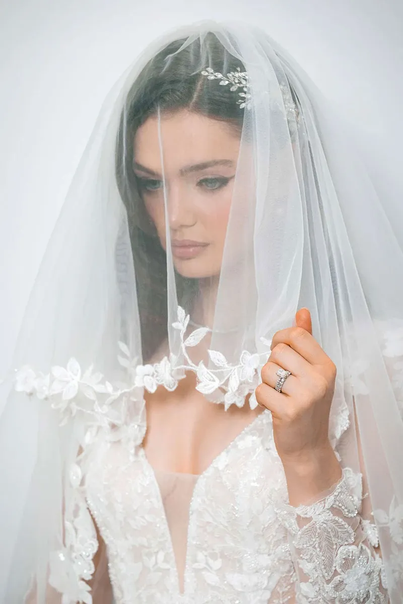 A bride, showcased by Tiara Studio under the Wedding Photography category, is seen in an elegant white lace wedding dress, delicately adorned with floral embroidery. She gazes softly downward, holding a sheer veil that gently drapes over her face, highlighting her subtle makeup and long, flowing hair. The intricate lace details and the serene expression capture the essence of elegance and refinement, ideal for wedding inspirations on Mariage Elegant, Canada.