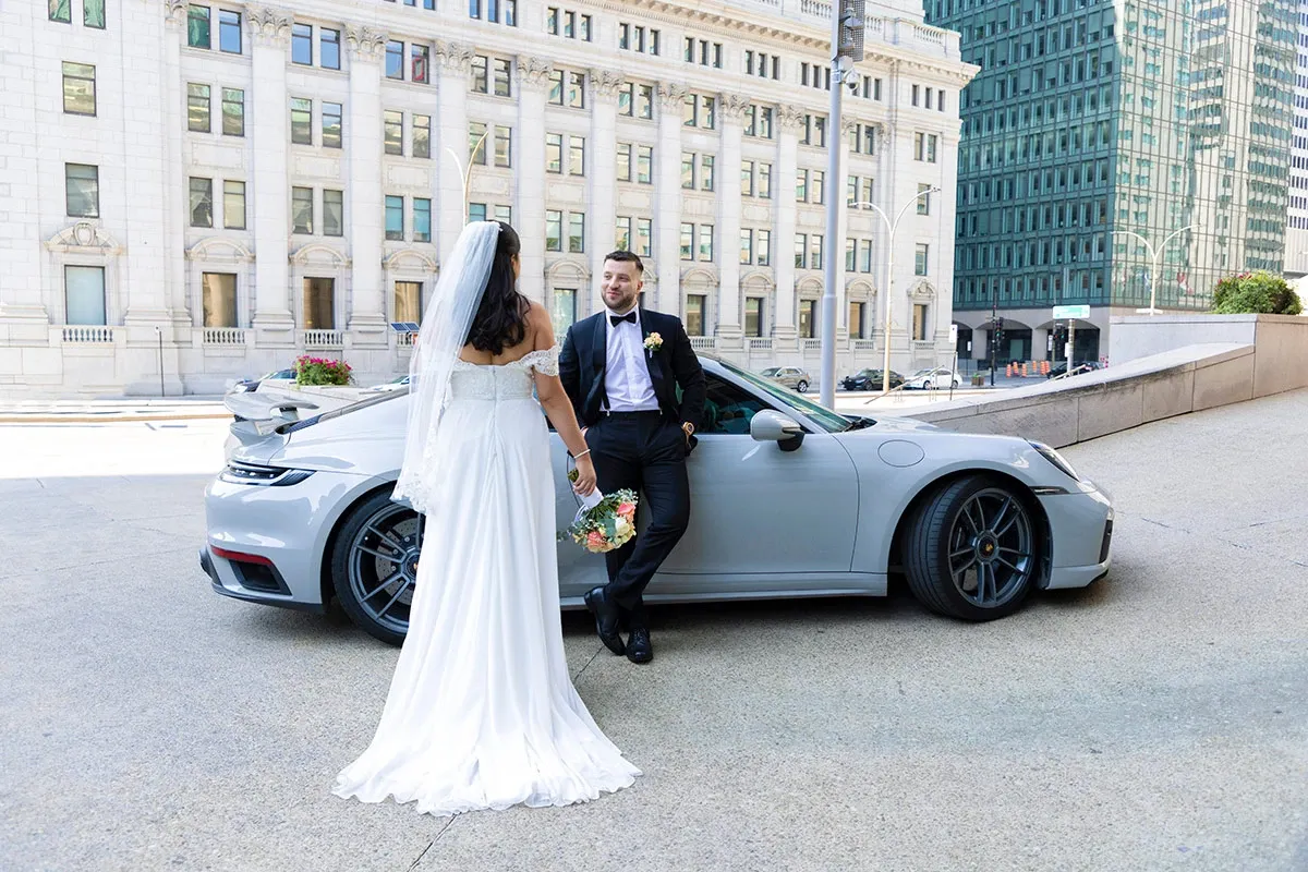 A bride and groom are captured in a candid moment by Tiara Studio, a renowned wedding photography vendor listed on Mariage Elegant in Canada. The groom, dressed in a sleek black tuxedo with a white shirt and bowtie, leans casually against a silver sports car parked on an urban street. The bride, wearing an elegant white gown and veil, holds a bouquet of flowers as she faces him. In the background, there is a grand building with tall windows and classical architecture, adding an urban sophistication to the wedding scene. This image beautifully showcases a modern couple surrounded by the bustling cityscape.