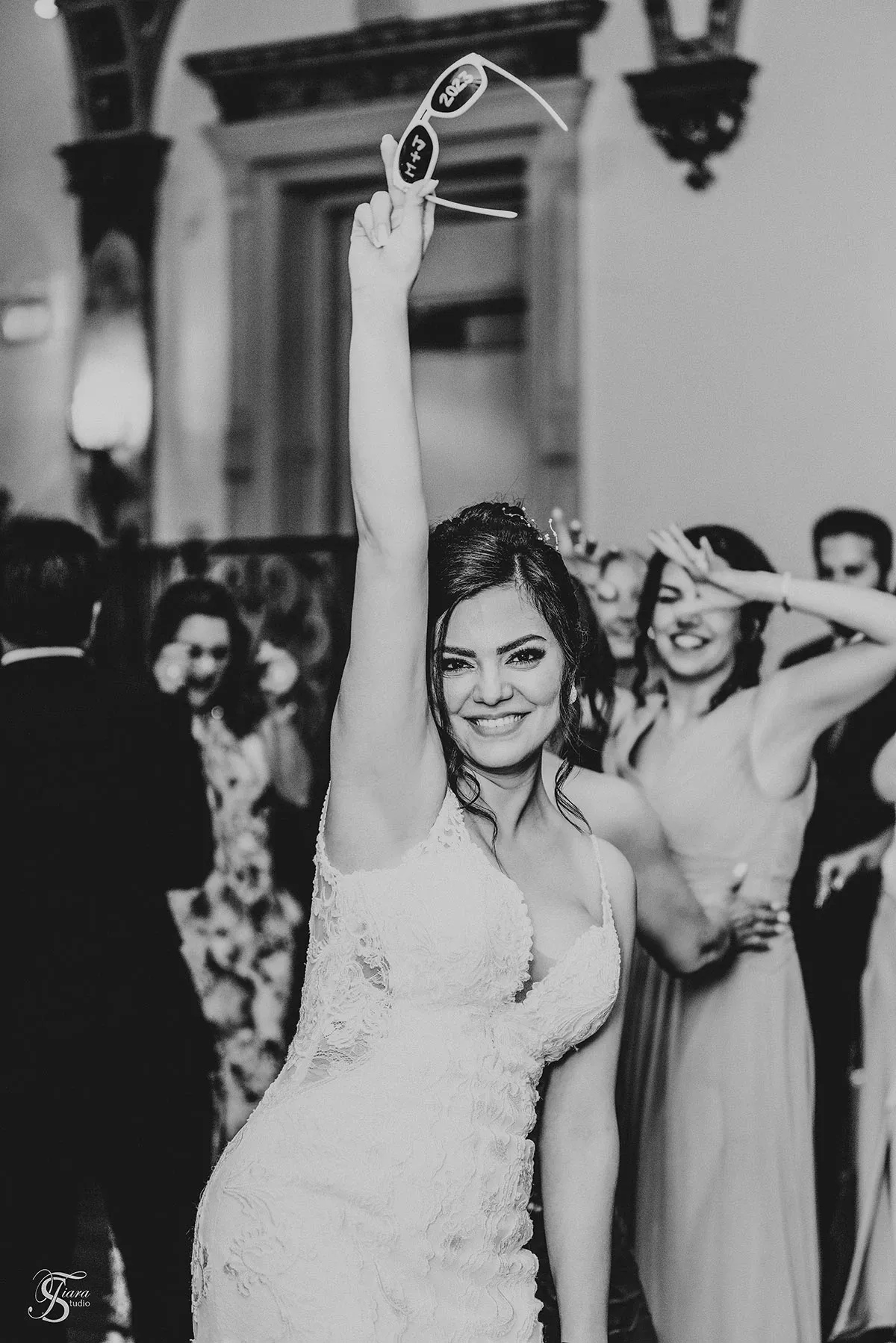 A joyful bride in a lace wedding dress from Tiara Studio is captured in a candid moment at her wedding reception, showcasing her excitement. She is holding a pair of sunglasses high in the air, surrounded by laughing guests in elegant attire. The black and white photograph, taken by a wedding photographer under the category of Wedding Photography, features an ornate venue with decorative arches in the background, adding to the celebratory atmosphere.