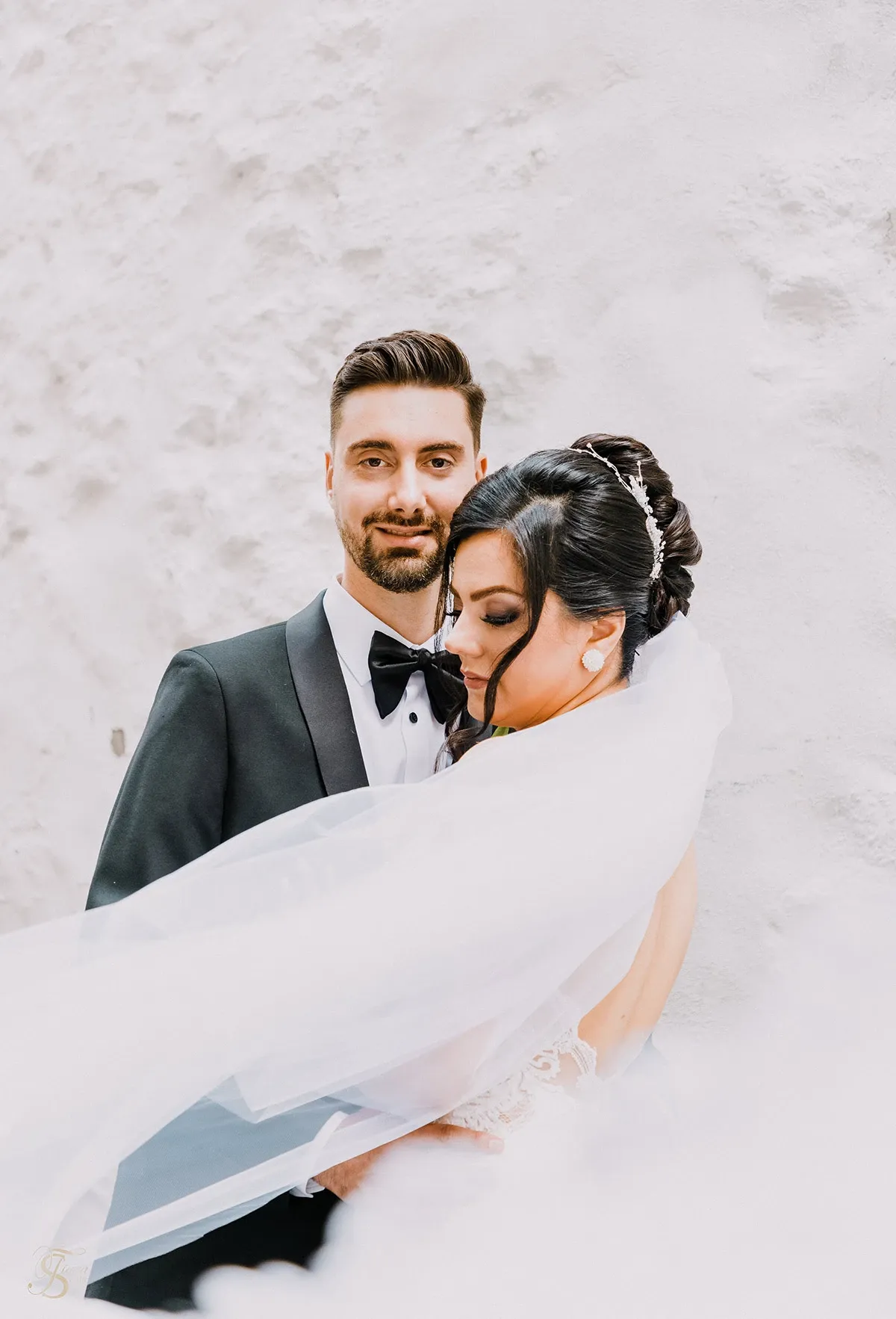 A bride and groom stand closely together against a textured white wall, exuding elegance and romance. The bride, wearing a beautiful lace wedding gown with a flowing veil, rests her head gently on the groom's shoulder. Her dark hair is styled in an intricate updo, adorned with a tiara, complementing her radiant appearance. The groom is dressed in a classic black tuxedo with a bow tie, looking directly at the camera with a composed expression. This image, captured by Tiara Studio under their Wedding Photography category, embodies the elegance and intimacy of the couple's special day, reflecting the sophisticated services offered by Mariage Elegant in Canada.