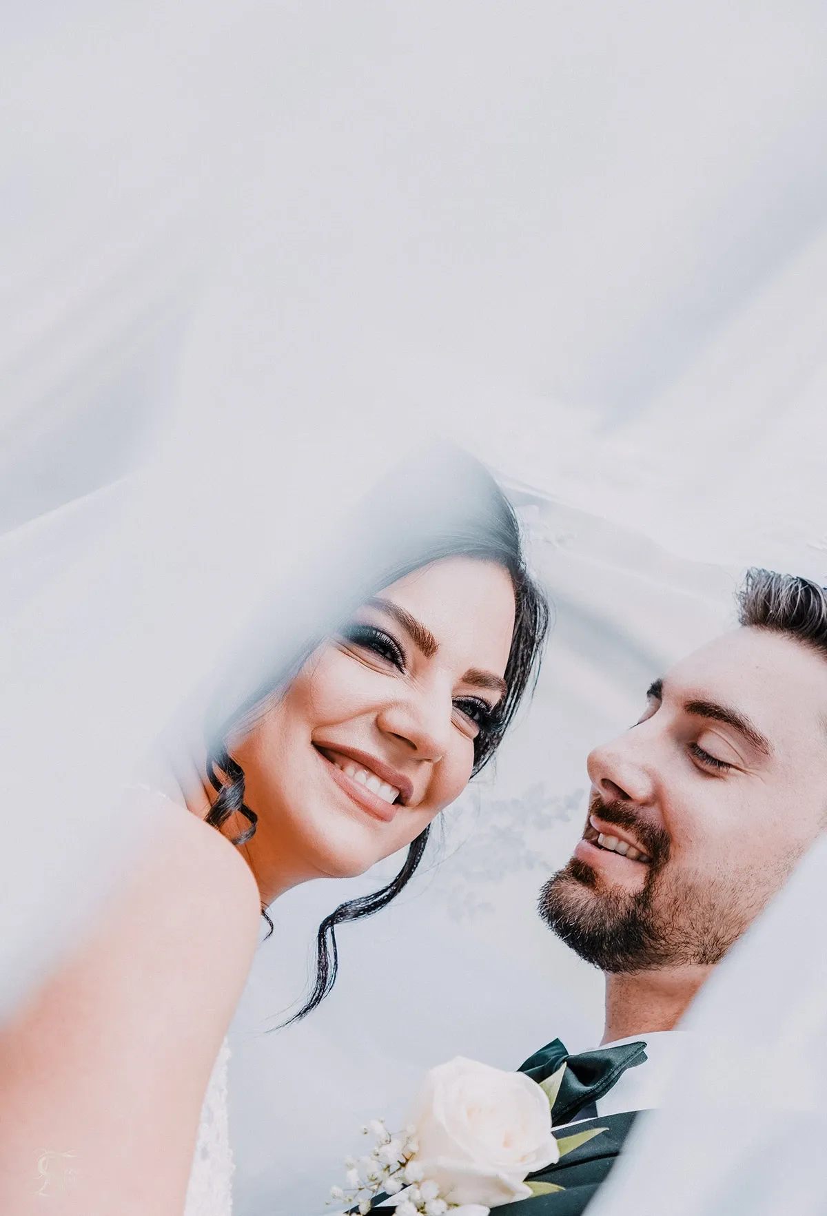 A joyful bride and groom are captured in a tender moment under a soft, flowing veil. The bride is smiling brightly, her dark hair elegantly styled, and she wears a delicate white dress. The groom, wearing a classic suit with a white rose boutonniere, gazes lovingly at her. This intimate and romantic scene is skillfully photographed by Tiara Studio, showcasing their expertise in wedding photography. The gentle draping of the veil creates a dreamy ambiance, emphasizing the couple's happiness on their special day. This image is featured on Mariage Elegant, a wedding planning and vendor listing site in Canada.