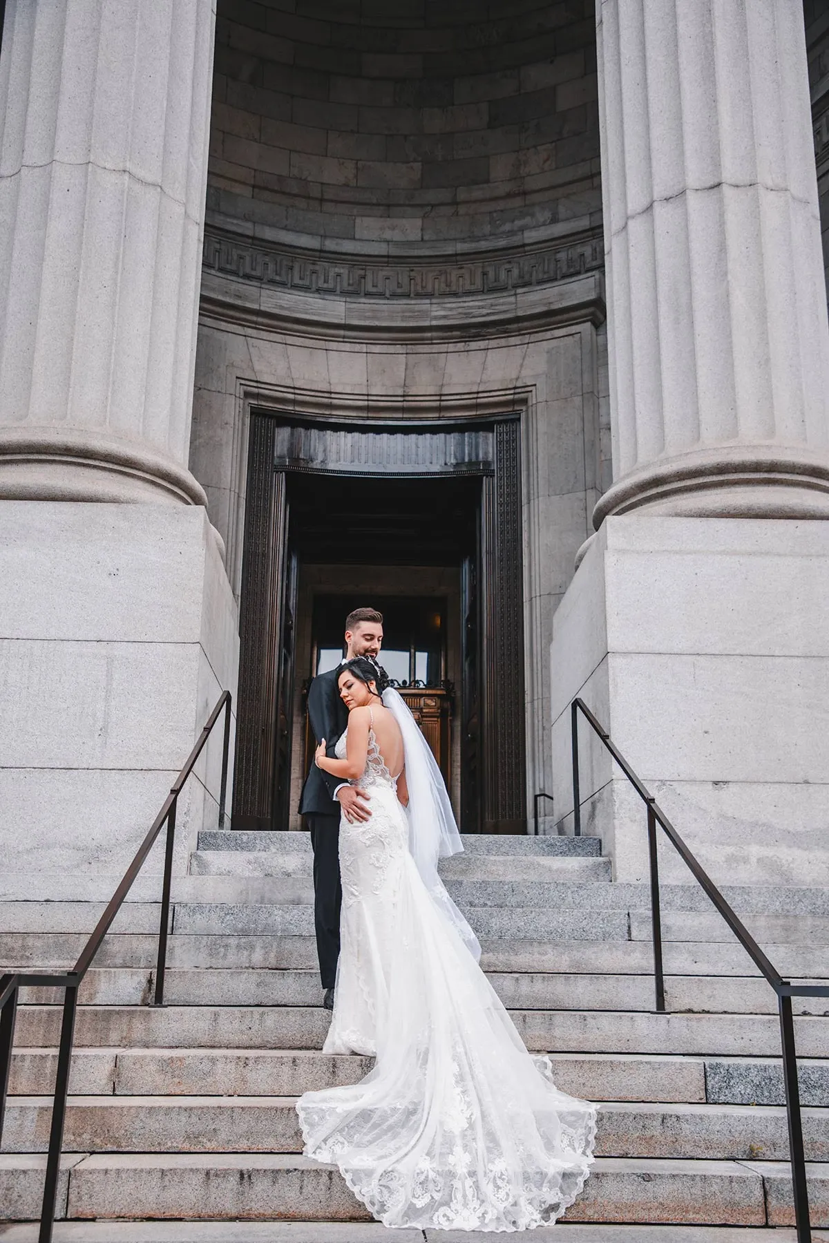 The image showcases a newlywed couple on the steps of an elegant stone building with grand columns, captured by Tiara Studio, a wedding photography vendor in the Mariage Elegant listing in Canada. The bride is wearing a stunning white lace gown with an open back, while the groom is dressed in a classic black tuxedo. They are embracing lovingly, creating a romantic and timeless scene. The backdrop of the historic architecture adds a touch of sophistication, making this wedding photograph a perfect representation of elegance and celebration.