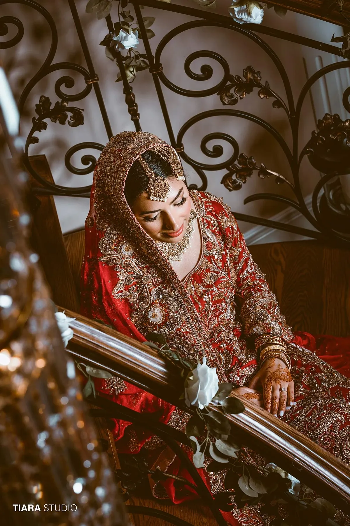 In this captivating image by Tiara Studio, a leading vendor in the Wedding Photography category on Mariage Elegant, Canada, a bride is elegantly seated, adorned in a stunning red and gold traditional bridal gown. Her ensemble is richly detailed with intricate embroidery and embellishments. She gracefully rests on a wooden bench next to an ornate wrought iron railing decorated with soft, white roses. The bride's hands feature beautiful mehndi designs, and she wears a shimmering gold headpiece, complementing her radiant makeup and serene expression. This photograph beautifully captures the essence of an elegant, traditional wedding moment.
