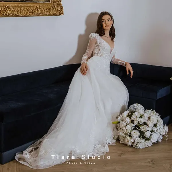A bride in an elegant white gown with lace sleeves and a flowing train poses gracefully on a dark blue sofa, her gaze directed thoughtfully. She is surrounded by an ornate gold picture frame on the wall and a large bouquet of white flowers placed on the floor. This image, captured by Tiara Studio, showcases their expertise in wedding photography, highlighting a moment of bridal poise and sophistication, perfect for those planning a wedding in Canada.