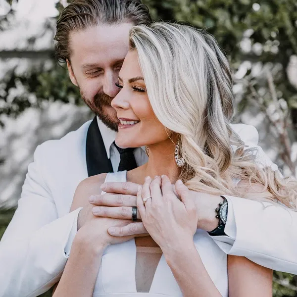 A joyful couple is captured on their wedding day by Tiara Studio, a vendor specializing in Wedding Photography. The groom, dressed in a stylish white suit, lovingly embraces the bride from behind. The bride, wearing a beautiful white gown, smiles radiantly. Her blonde hair cascades in soft waves, and she gently holds the groom’s hands over her chest. The background features lush greenery, adding a natural and elegant touch to the moment. This image portrays the artistry of Tiara Studio in capturing intimate and heartwarming wedding moments.