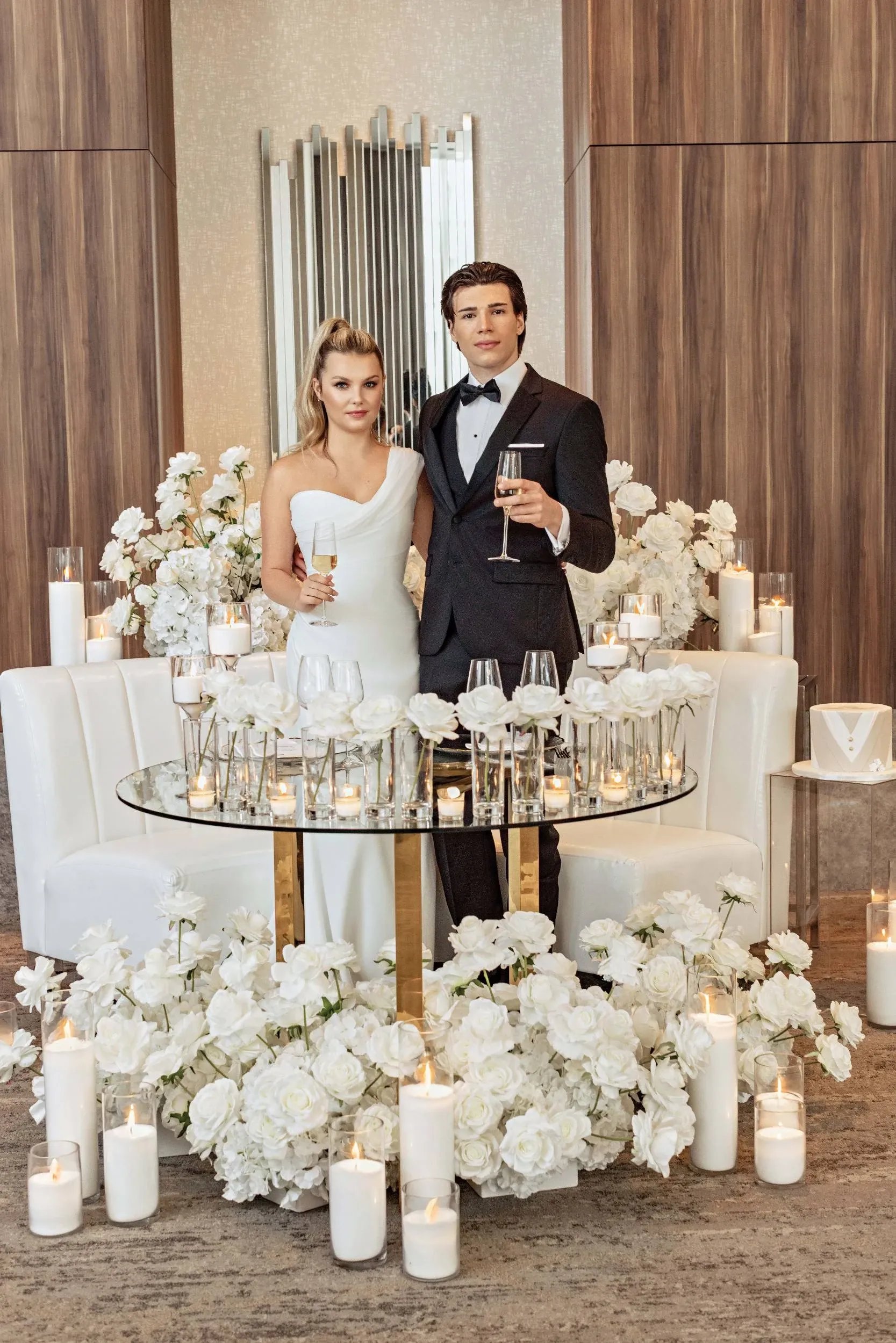 A couple elegantly dressed in wedding attire stands in the Westbury Ballroom, a featured wedding venue on Mariage Elegant, Canada. The bride wears a stylish one-shoulder white gown, and the groom is in a classic black tuxedo. They are positioned in front of a sophisticated glass table adorned with several wine glasses and surrounded by abundant white floral arrangements and lit candles, creating a romantic and luxurious ambiance. The setting reflects the venue's commitment to elegance and grandeur, ideal for weddings.