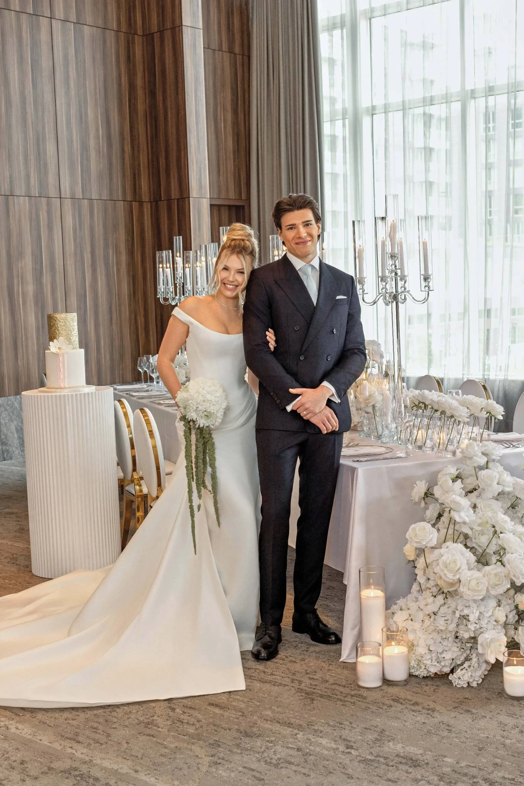 A bride and groom stand elegantly in the Westbury Ballroom, a stunning wedding venue in Canada, featured on Mariage Elegant. The bride is wearing a classic white wedding gown and holding a bouquet of white flowers, while the groom is dressed in a sharp dark suit. They are posing beside a beautifully set wedding table adorned with chic white floral arrangements, tall silver candelabras, and glowing candles. A stylish white wedding cake is displayed on a nearby pedestal. The ballroom features high ceilings and large windows, allowing natural light to illuminate the sophisticated setting.