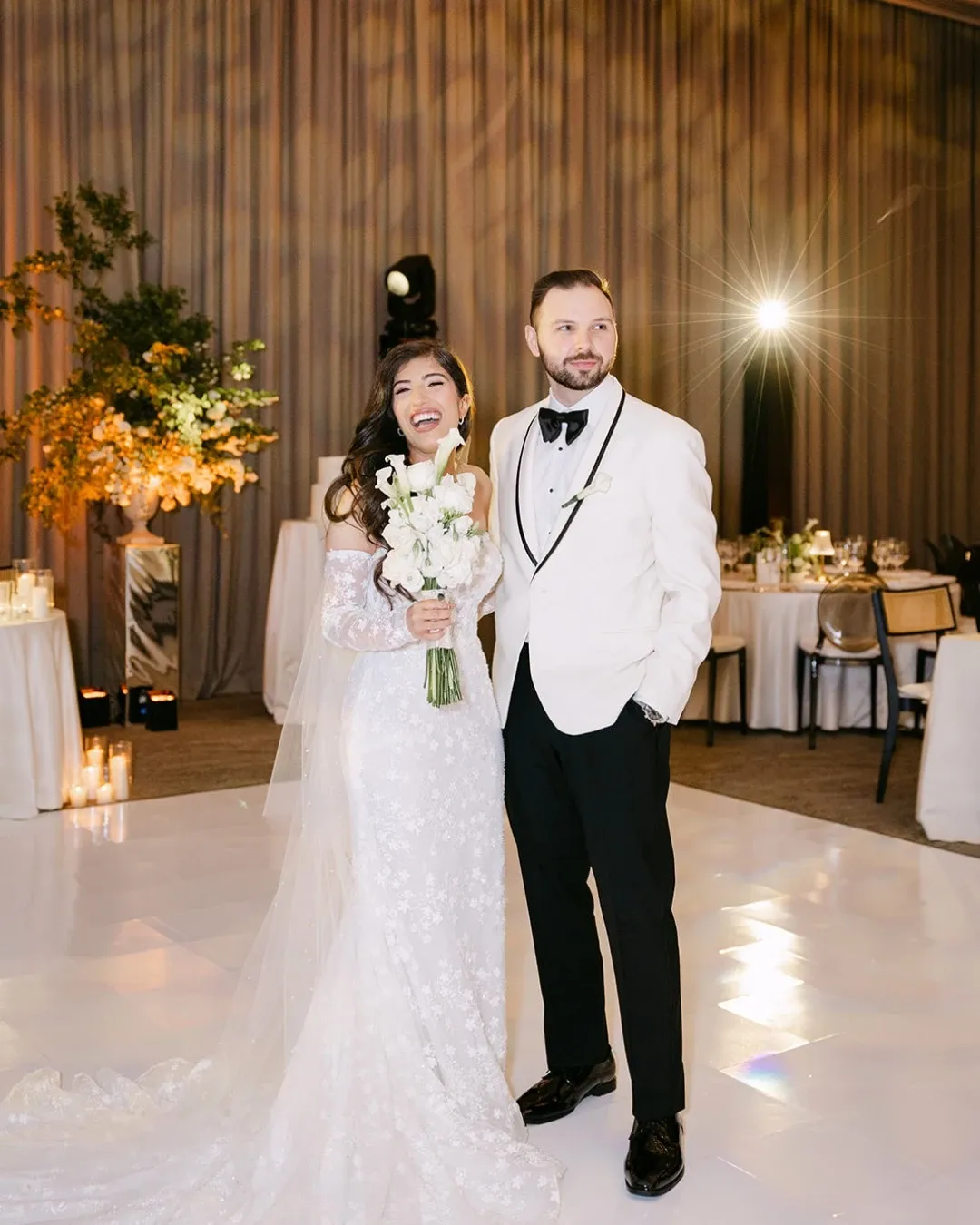 A bride and groom stand smiling on a pristine white dance floor inside the Westbury Ballroom, a premier wedding venue in Canada, part of Mariage Elegant's listings. The bride is wearing an elegant lace wedding gown with a long train and holding a bouquet of white flowers. The groom is dressed in a classic white tuxedo jacket with a black bow tie. Soft, ambient lighting and tall floral arrangements with greenery provide a romantic backdrop, while tables adorned with candles and elegant table settings are visible in the background, accentuating the upscale atmosphere of the venue.