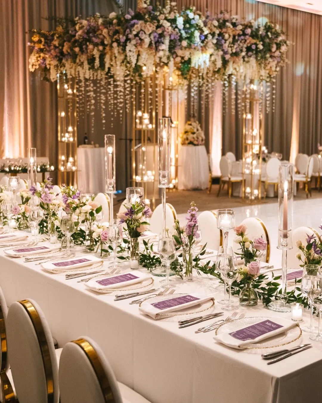 This image showcases an elegantly decorated wedding venue at Westbury Ballroom, categorized under Wedding Venues on the Mariage Elegant, Canada site. The setting features a beautifully arranged long banquet table adorned with white tablecloths, surrounded by white cushioned chairs with gold detailing. Each place setting includes fine china, glassware, and lavender-colored napkins, adding a touch of sophistication. The table is artistically decorated with floral arrangements consisting of soft purple, pink, and white flowers, complemented by greenery. Tall, slender candle holders with white candles line the center of the table, providing a romantic ambiance. In the background, there are luxurious floral installations hanging from the ceiling, featuring cascading greenery and flowers, enhancing the elegant atmosphere of the ballroom.