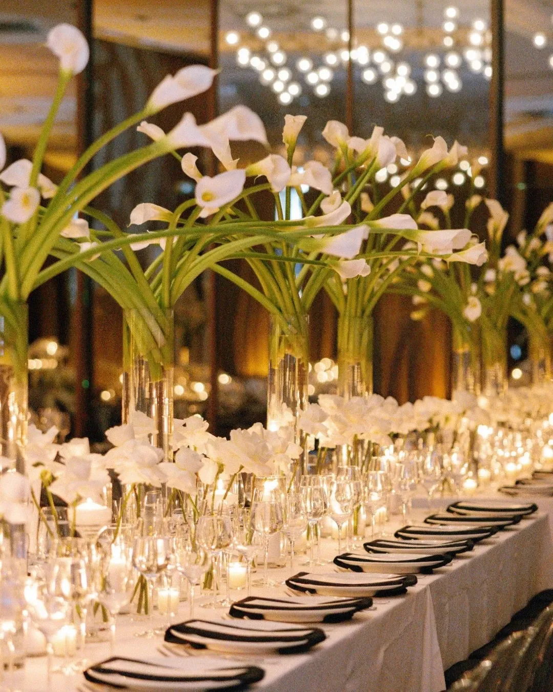 The image showcases an elegantly decorated long dining table at Westbury Ballroom, categorized under wedding venues. The table is adorned with tall, clear vases holding large, graceful calla lilies that create a stunning visual effect. Soft candlelight enhances the romantic atmosphere, reflecting off the sleek glassware and polished cutlery set neatly on the white tablecloth. This setting exemplifies the sophisticated ambiance ideal for weddings at this venue.