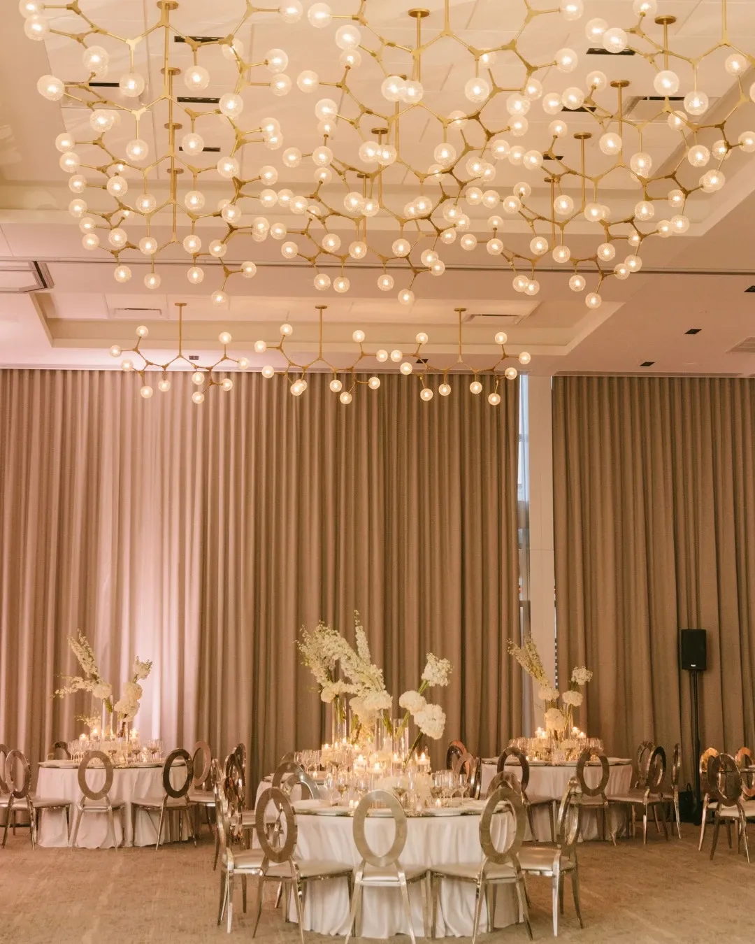 The image showcases the Westbury Ballroom, a premier wedding venue under the Mariage Elegant vendor listing in Canada. The ballroom features an elegant setting with opulent chandeliers hanging from the ceiling, casting a warm, inviting glow over the space. Round tables are elegantly arranged with white tablecloths and decorated with tall floral centerpieces, enhancing the sophisticated atmosphere. The soft beige curtains and subtle lighting contribute to the venue's luxurious ambiance, making it an ideal setting for memorable wedding celebrations.