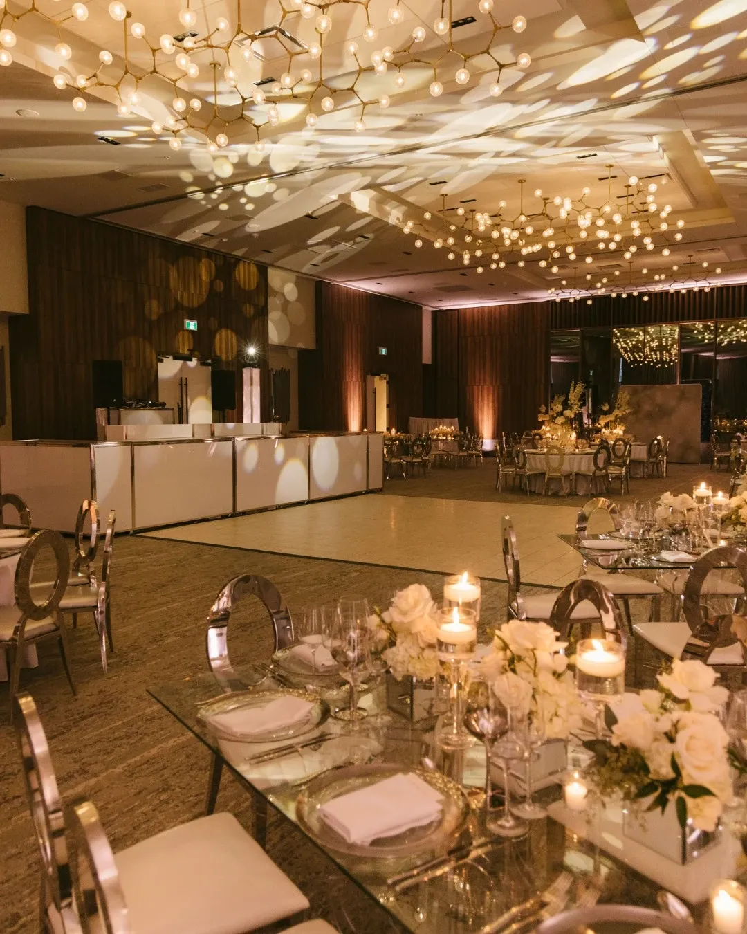The image showcases the Westbury Ballroom, a wedding venue under the category of Wedding Venues for Mariage Elegant, Canada. The ballroom is elegantly decorated with intricate light fixtures casting warm patterns on the ceiling. Round and rectangular tables are beautifully set with white linens, crystal wine glasses, and floral centerpieces composed of white roses and candles. The dance floor is spacious and surrounded by stylish chairs with metallic finishes. The ambiance of the venue is sophisticated and inviting, ideal for hosting a luxurious wedding reception.