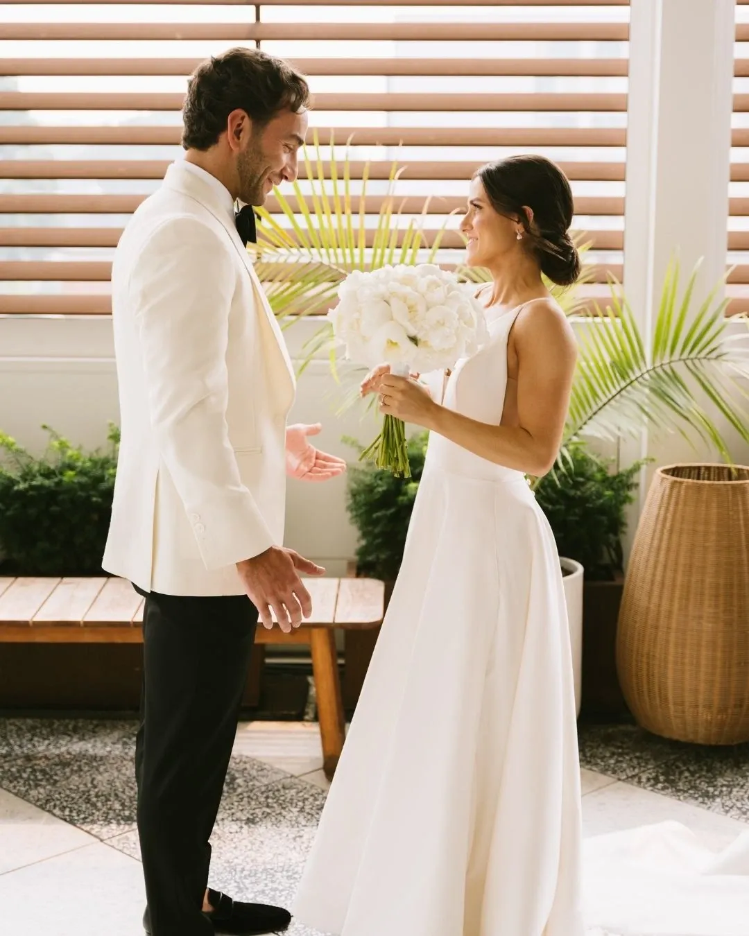 A bride and groom share a joyful moment at the Westbury Ballroom, a premier wedding venue in Canada. The bride is elegantly dressed in a flowing white gown and holds a bouquet of white flowers. The groom wears a classic white tuxedo jacket paired with black pants. Behind them, potted plants and wooden blinds add a natural and sophisticated touch, enhancing the intimate ambiance of the wedding setting at the Westbury Ballroom.