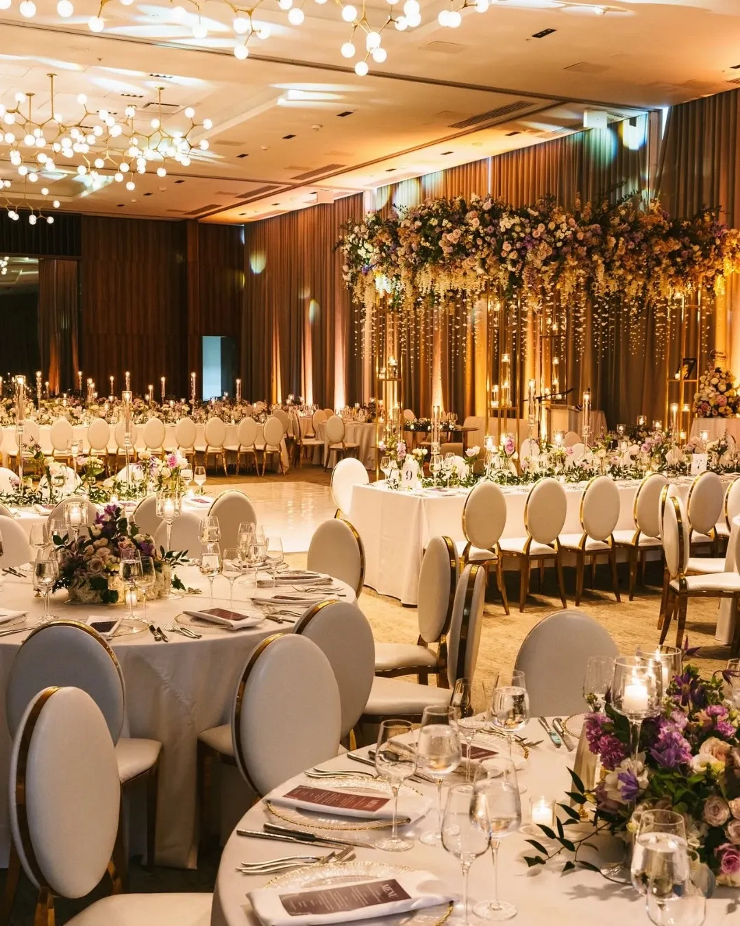 The image showcases an elegant wedding setup at Westbury Ballroom, categorized as a wedding venue on Mariage Elegant, Canada. The ballroom features beautifully arranged round tables adorned with white tablecloths and delicate floral centerpieces in shades of pink and purple. Exquisite gold-rimmed chairs complement the luxurious decor. A magnificent floral arch adorned with cascading flowers and greenery is prominently displayed in the background, alongside an array of lit candles that create a warm, inviting atmosphere. Overhead, a stunning display of modern chandeliers illuminates the room, enhancing the sophisticated ambiance ideal for a wedding celebration.